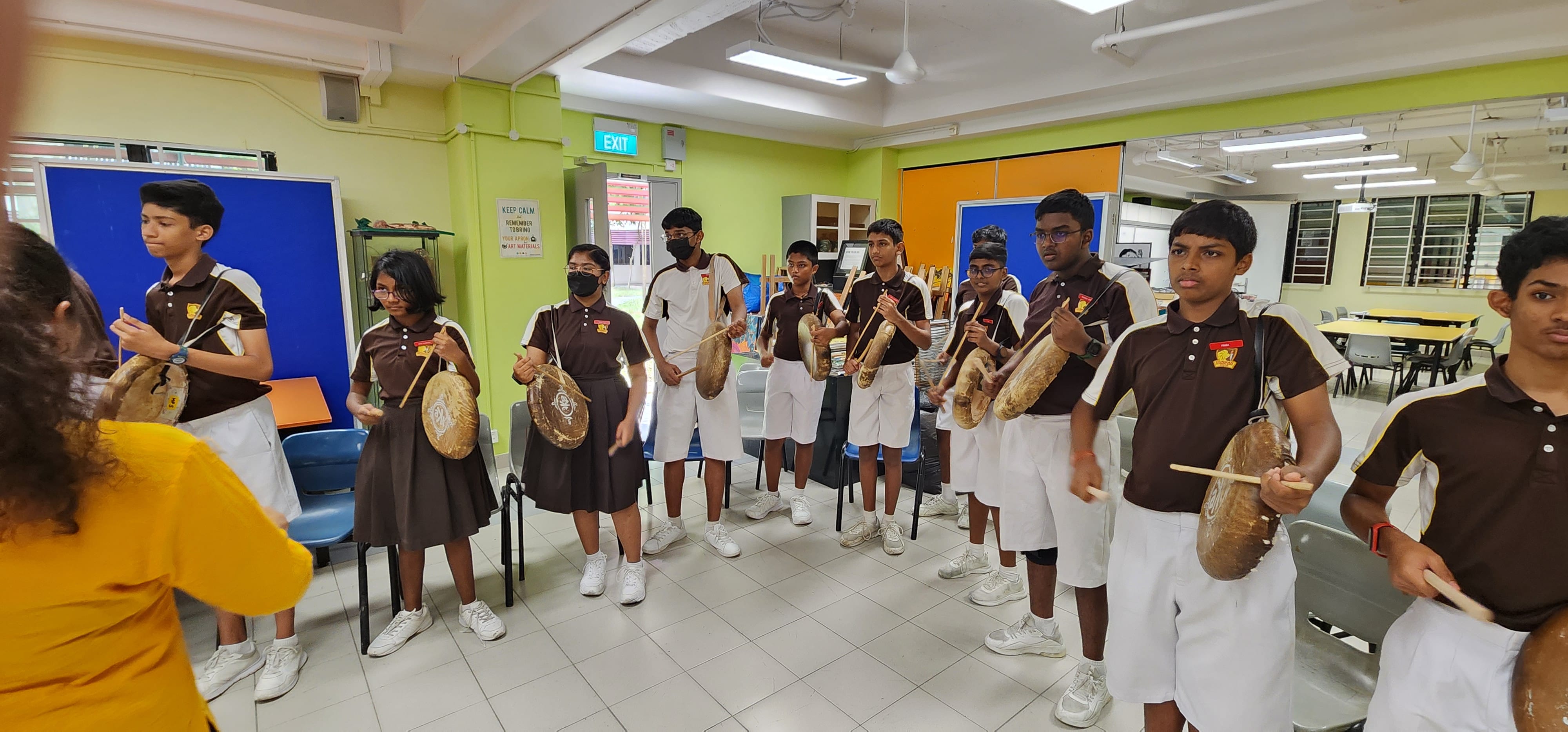 Students in brown uniforms stand in a row indoors, each holding a round hand drum and striking it with a stick.