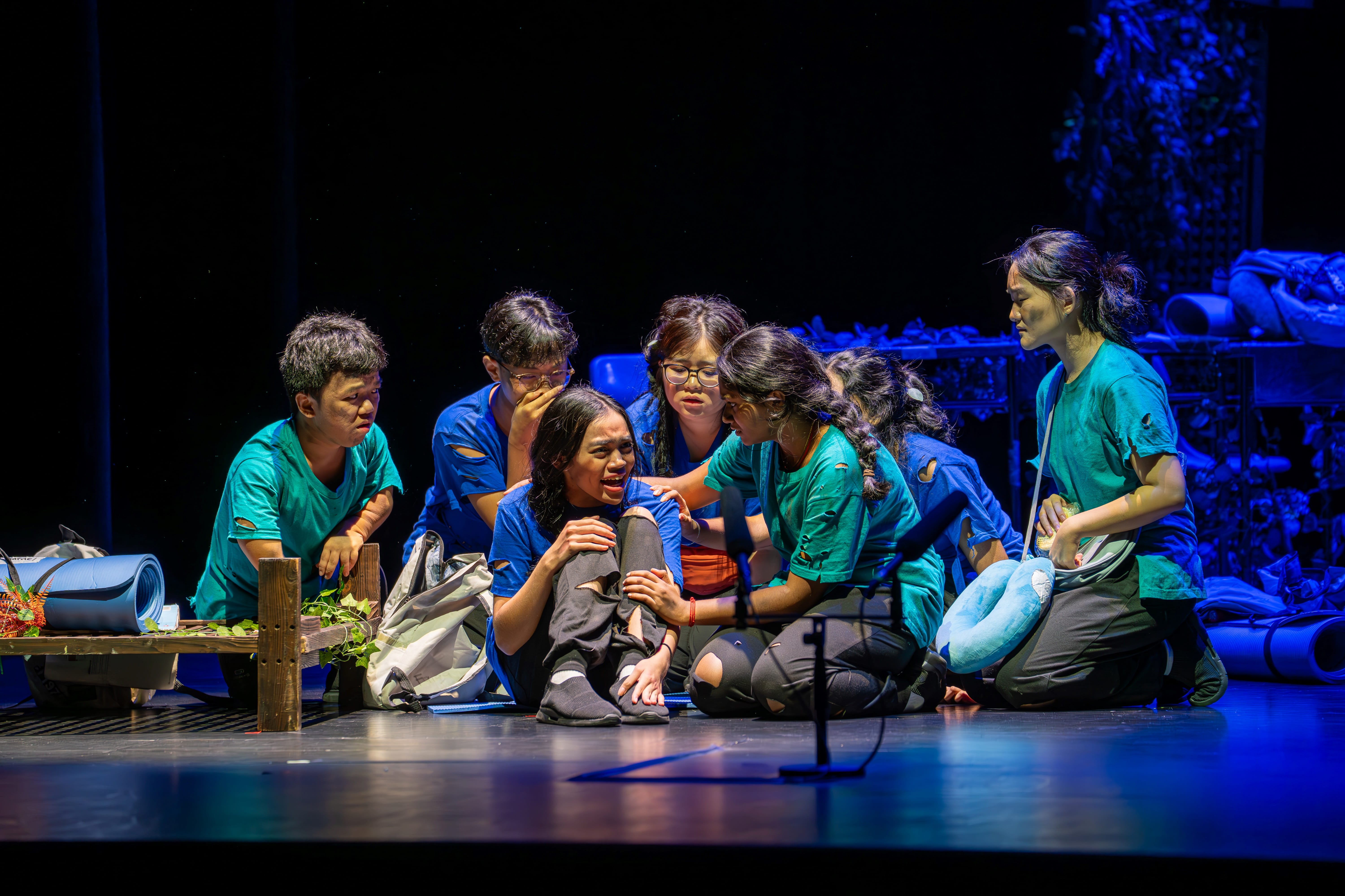 A group in blue and green shirts kneel onstage, comforting a crying seated woman under blue lighting.