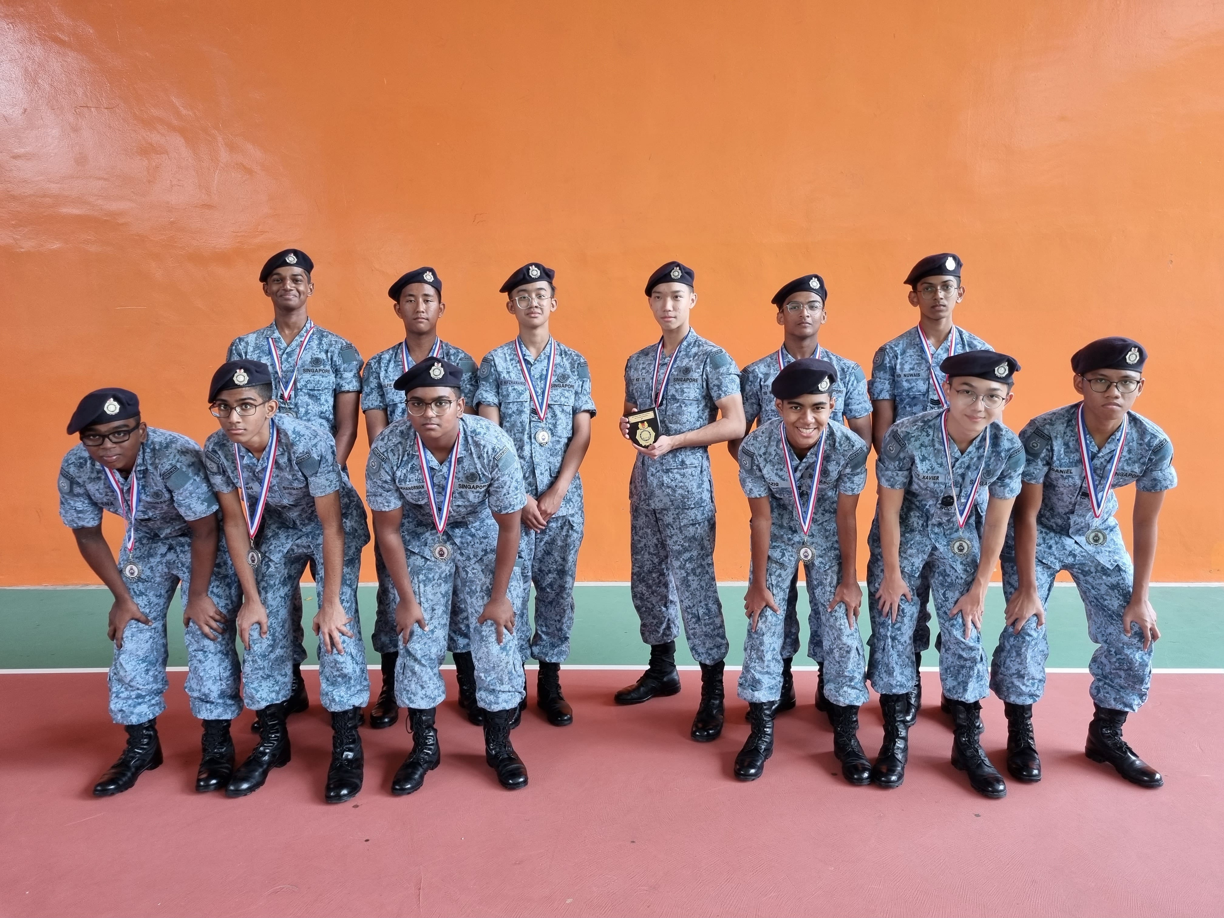 Twelve youths in blue camo uniforms and berets pose with medals; one holds a trophy before an orange wall.