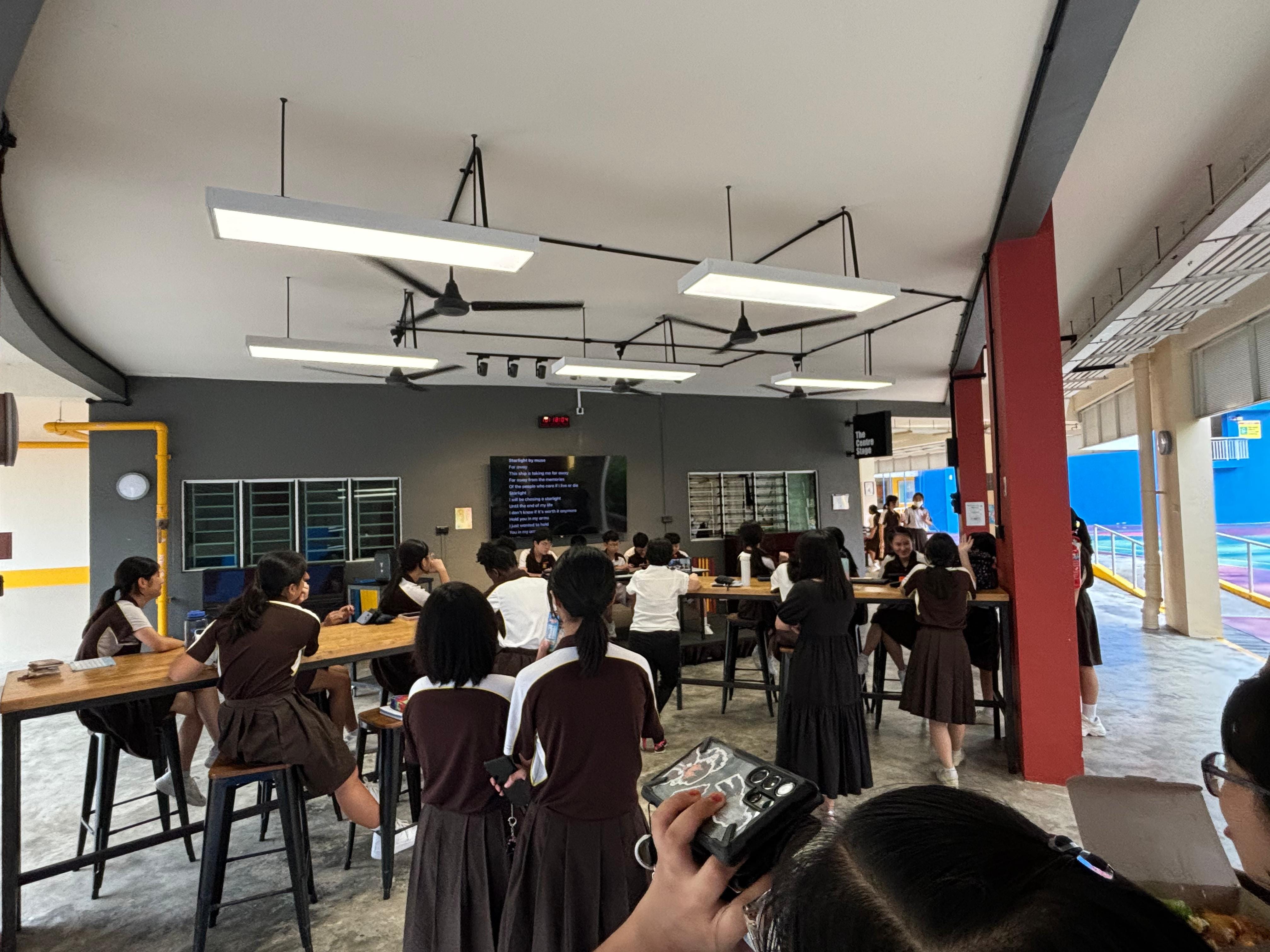 Students in dark uniforms sit and stand at tall wooden tables in an indoor space with ceiling fans and a screen on the wall.