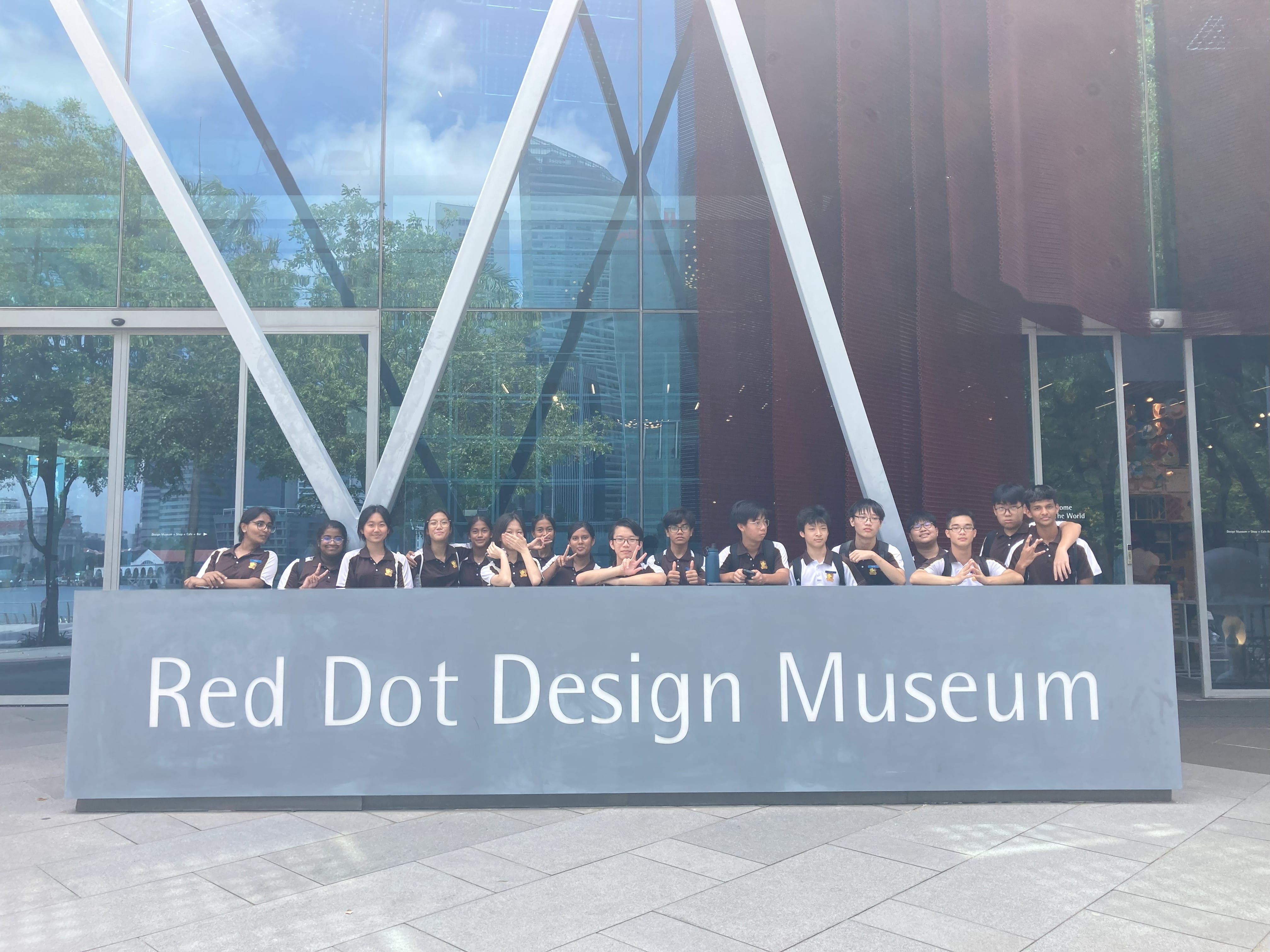 A group of students in brown uniforms posing behind the Red Dot Design Museum sign outdoors.