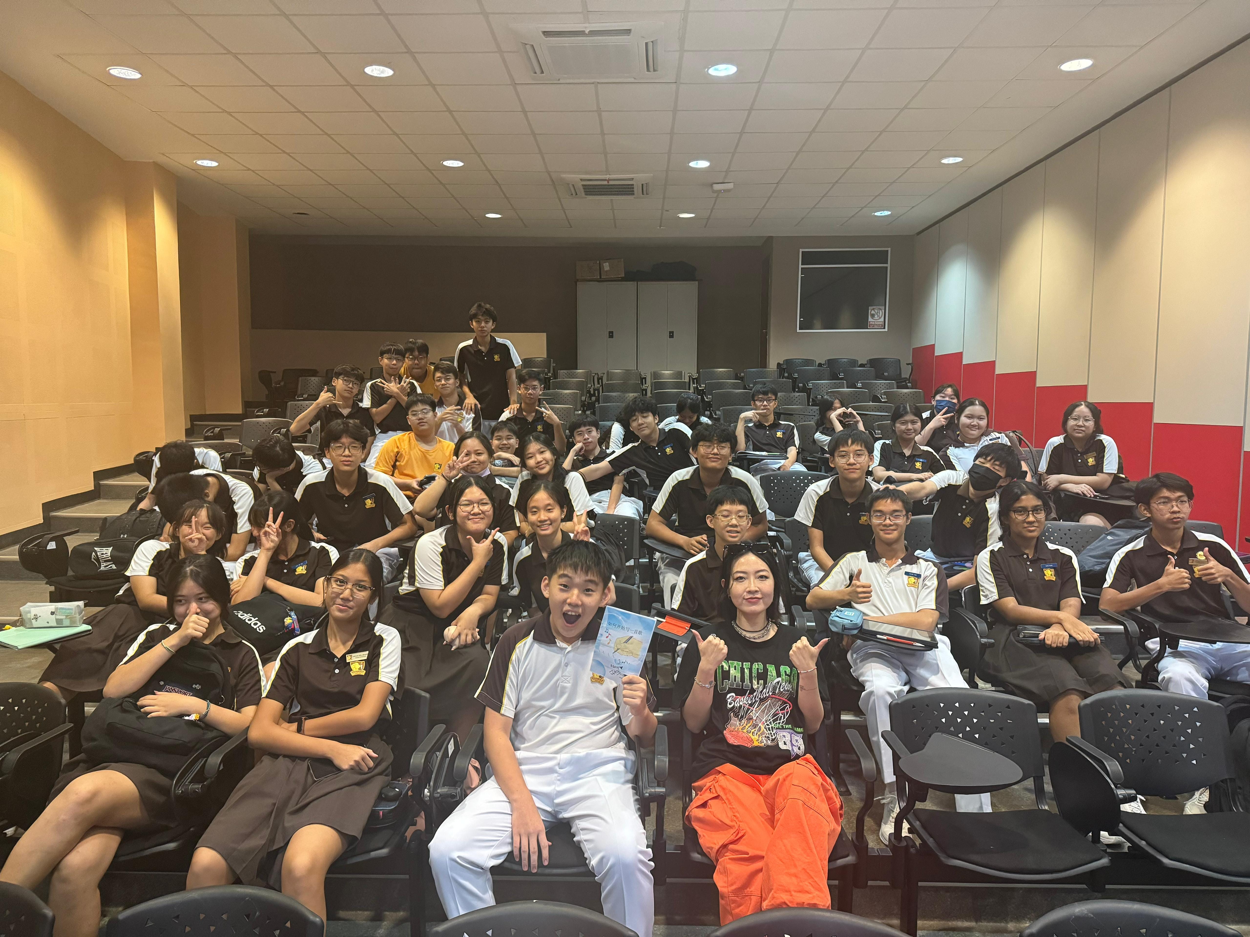 A large group of students in brown and white uniforms pose and smile in a tiered lecture hall.