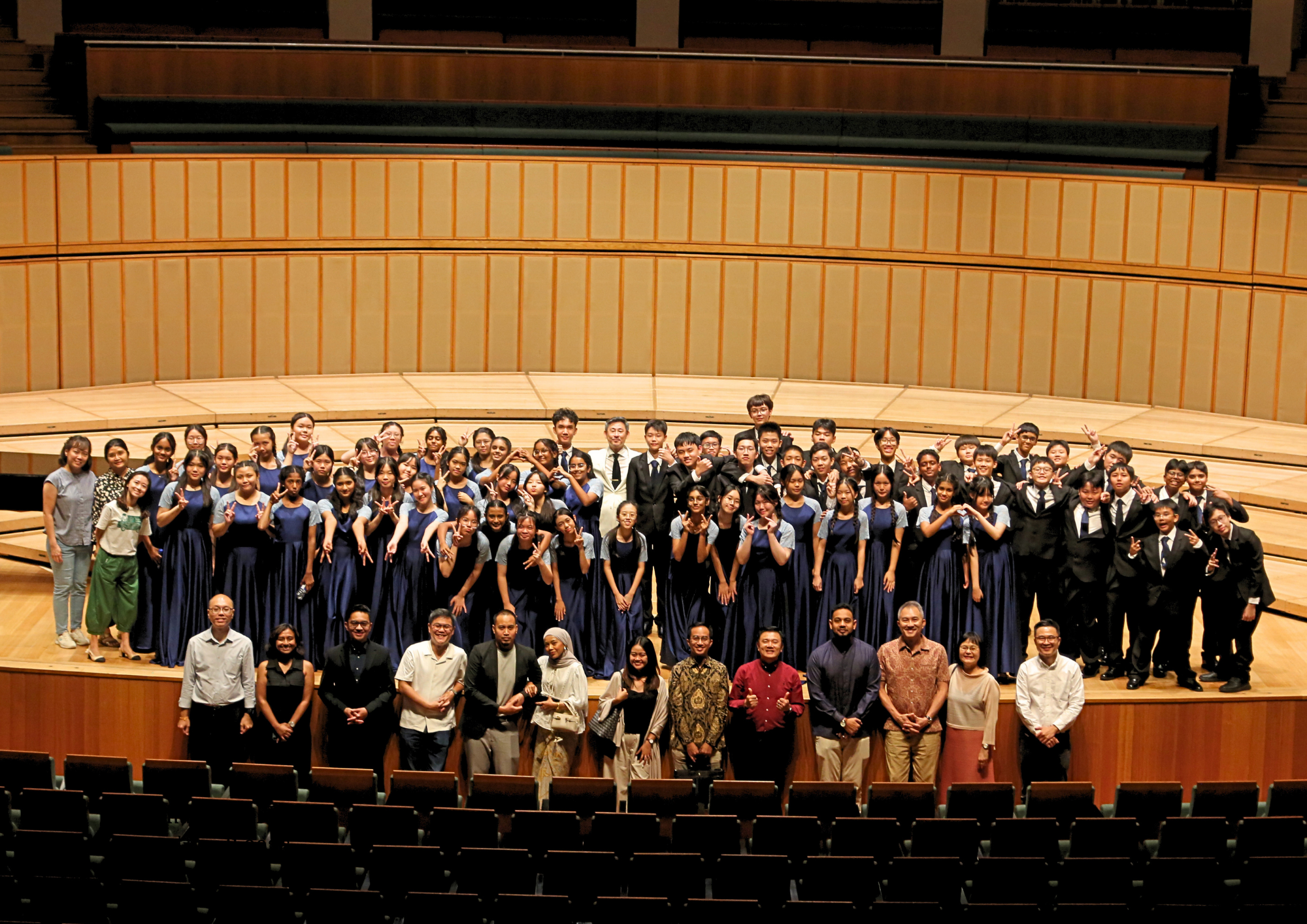 Large group on concert hall stage; many in navy dresses and black suits, some making peace signs