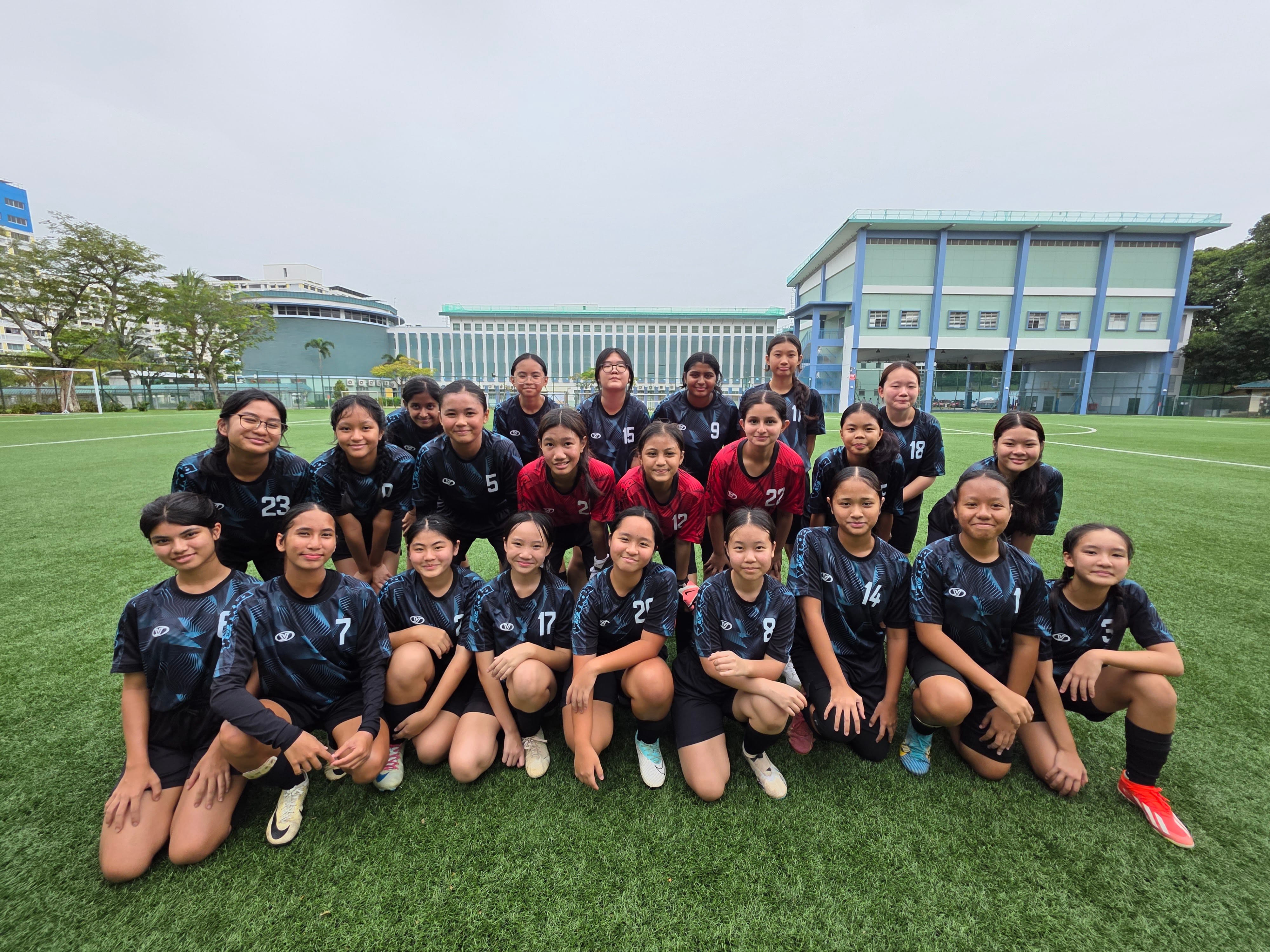 A girls soccer team poses in three rows on a green field, most wearing dark numbered jerseys, a few in red.