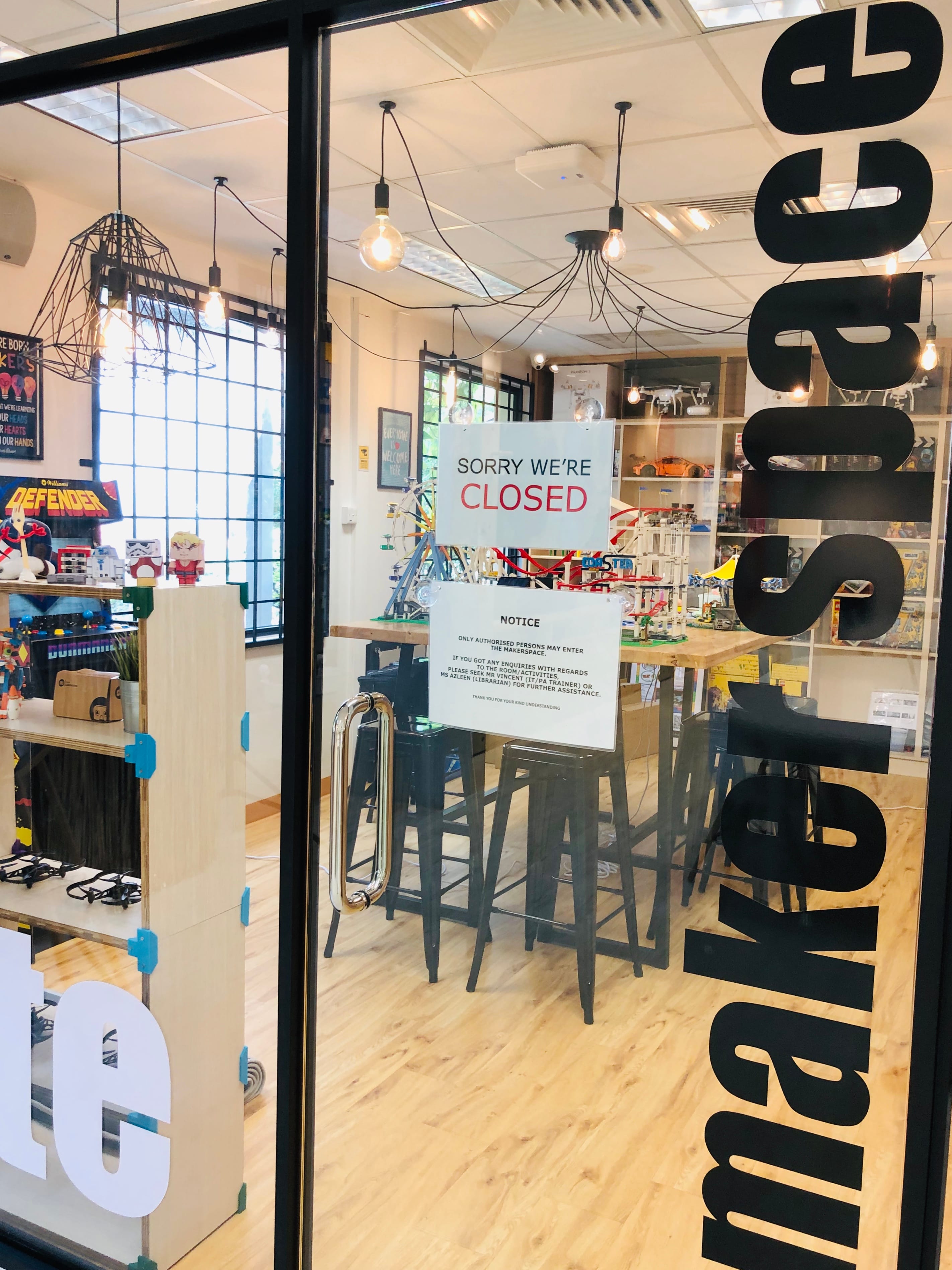 Glass door of a makerspace with a "Sorry We're Closed" sign, showing shelves with toys and bar stools inside.