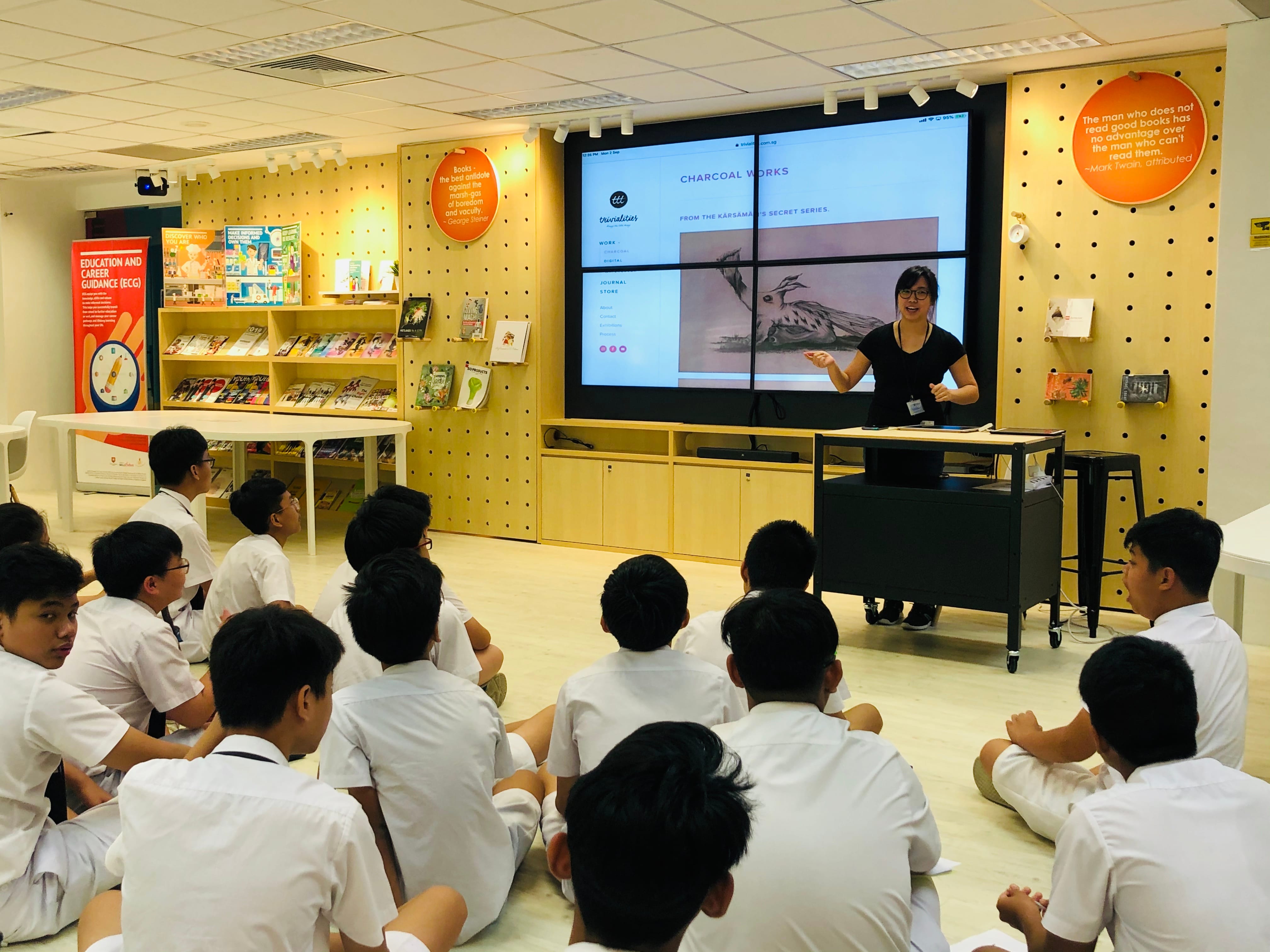 A woman presents to students in white uniforms sitting on the floor in a library, with a screen showing "Charcoal Works."