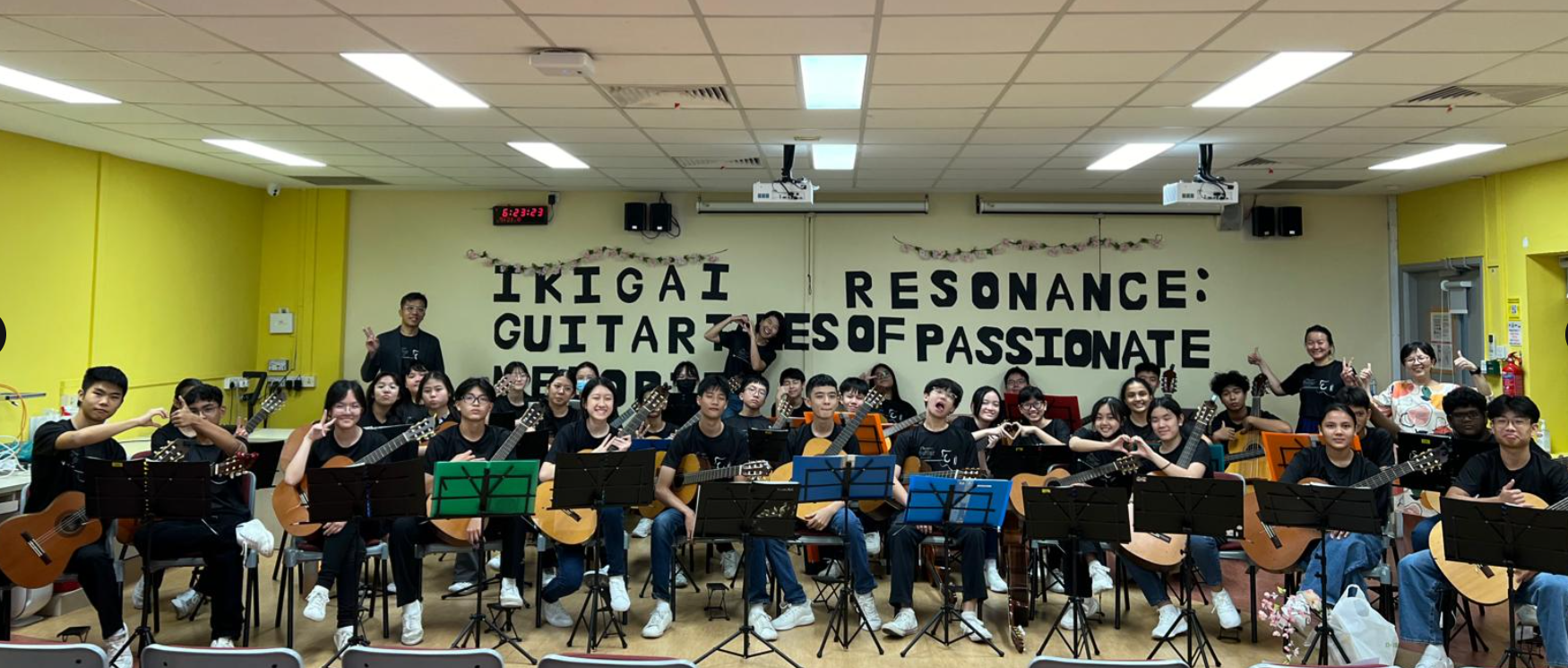 A large group of students in black clothes holding guitars, seated before a wall reading "Ikigai Resonance: Guitar Notes of Passionate."