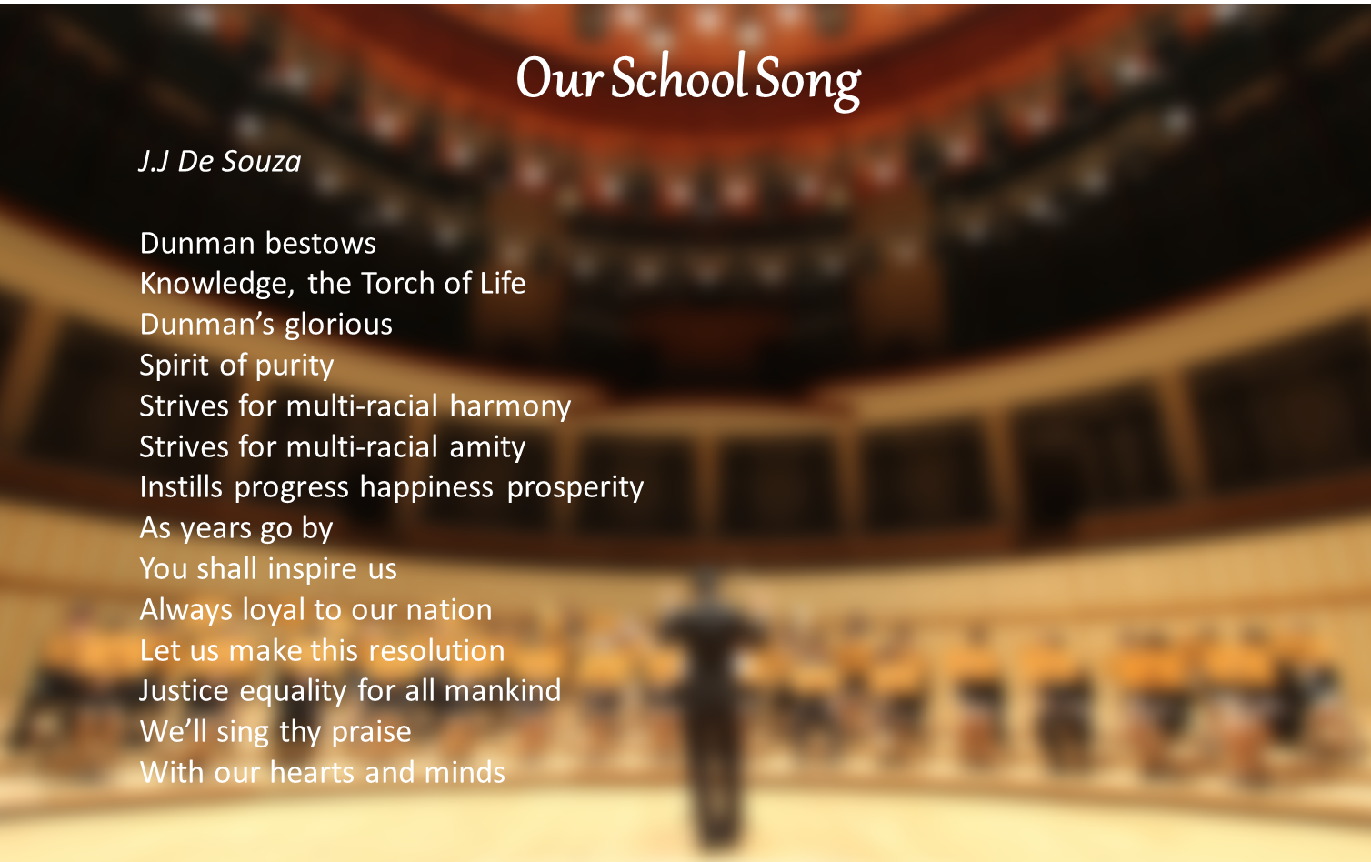 Slide titled Our School Song by J.J De Souza displaying full song lyrics over a blurred concert hall background.