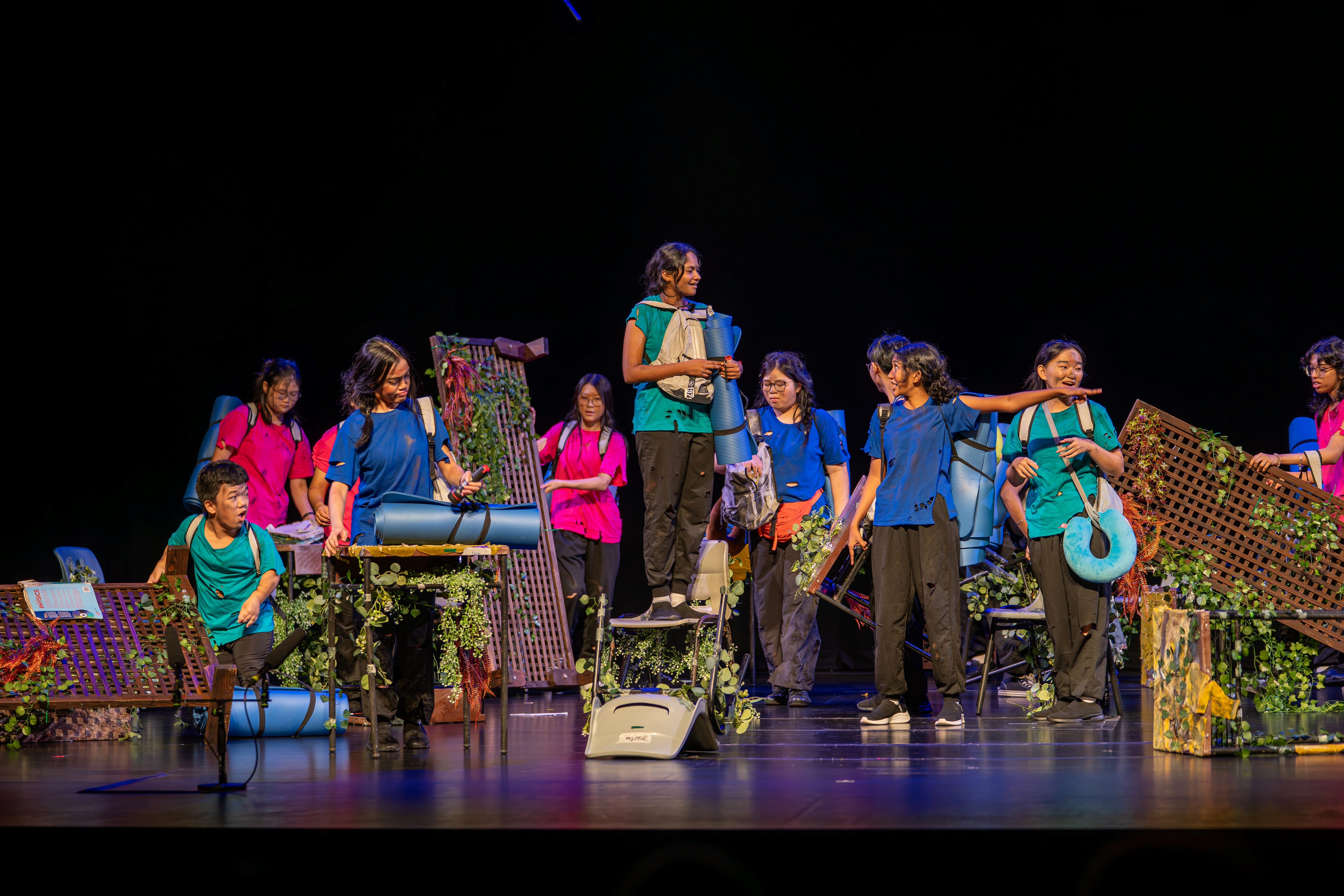 Group of performers onstage in colorful shirts among chairs, vines, and large props, one pointing right.
