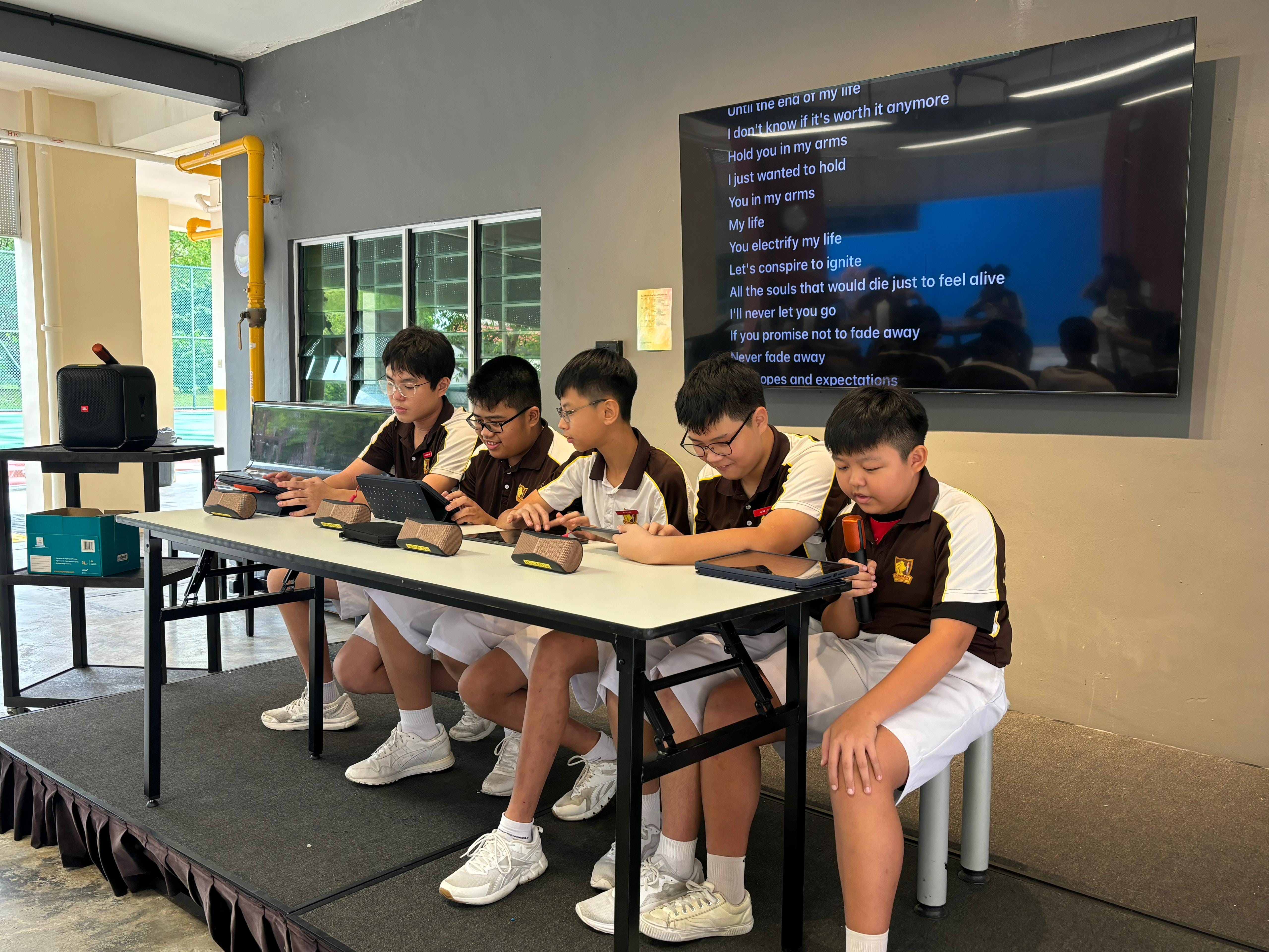 Five boys in school uniforms sit at a table with tablets on a stage, with song lyrics displayed on a screen behind them.