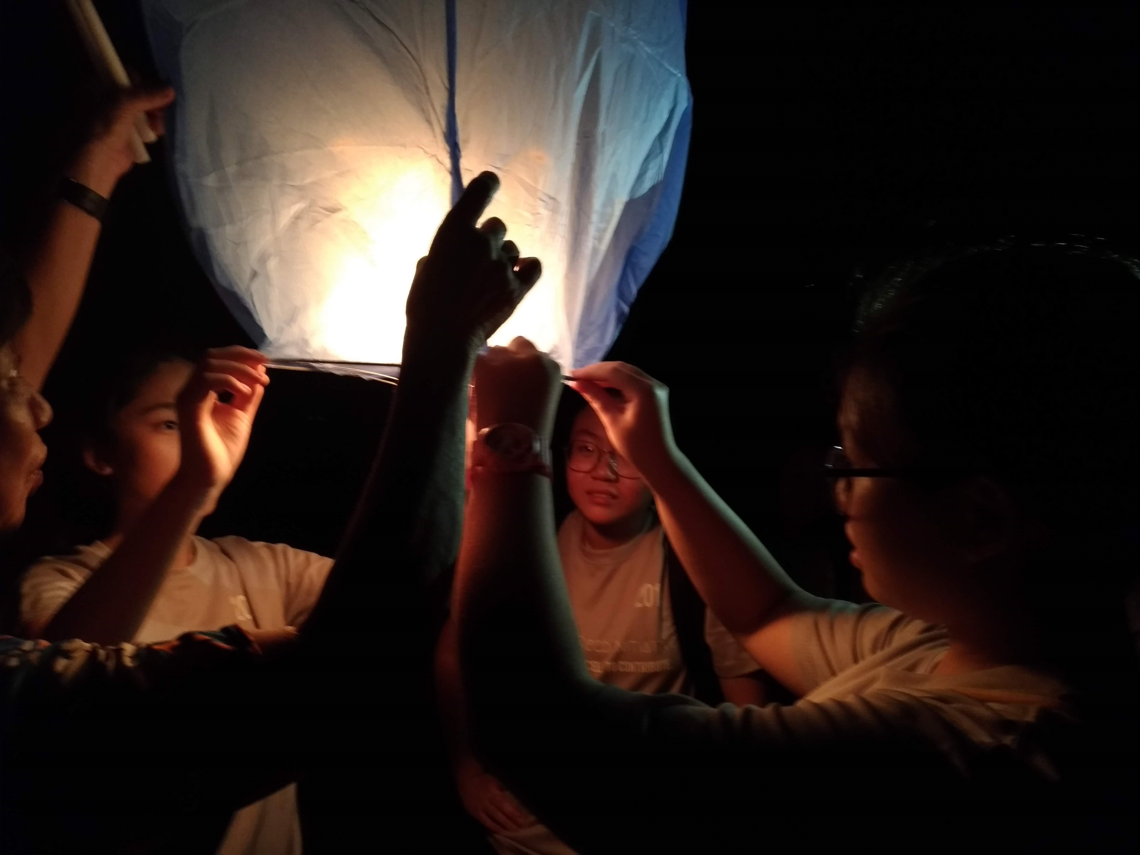 Several people hold up a glowing white and blue sky lantern together at night.