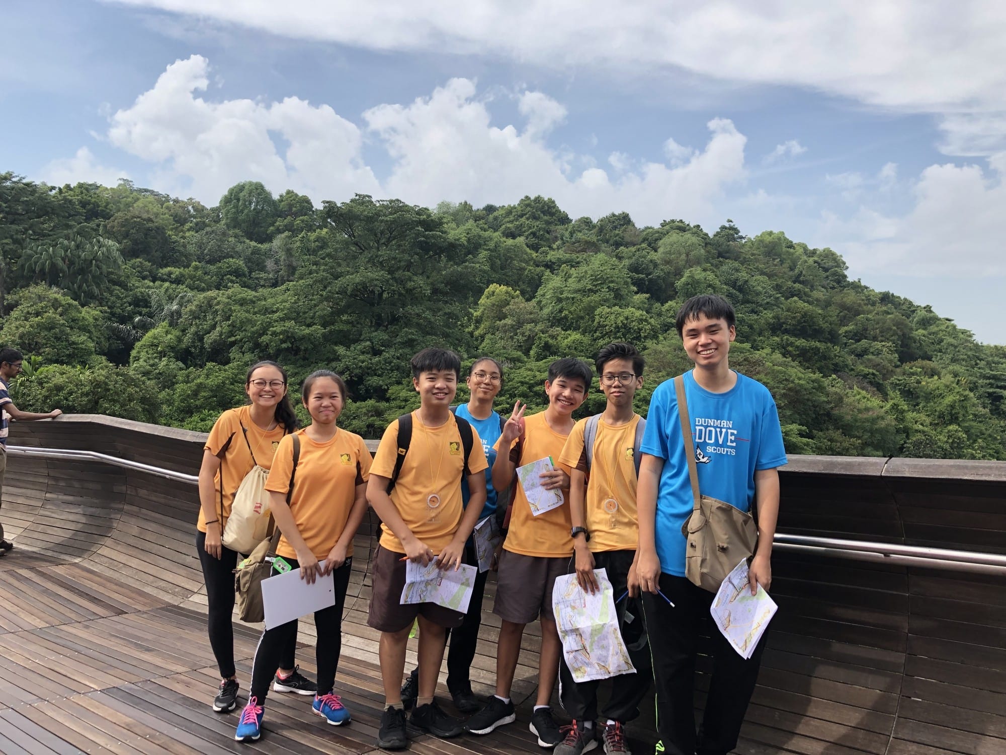Seven young students in orange and blue scout shirts hold maps on a wooden deck overlooking a green forested hillside.