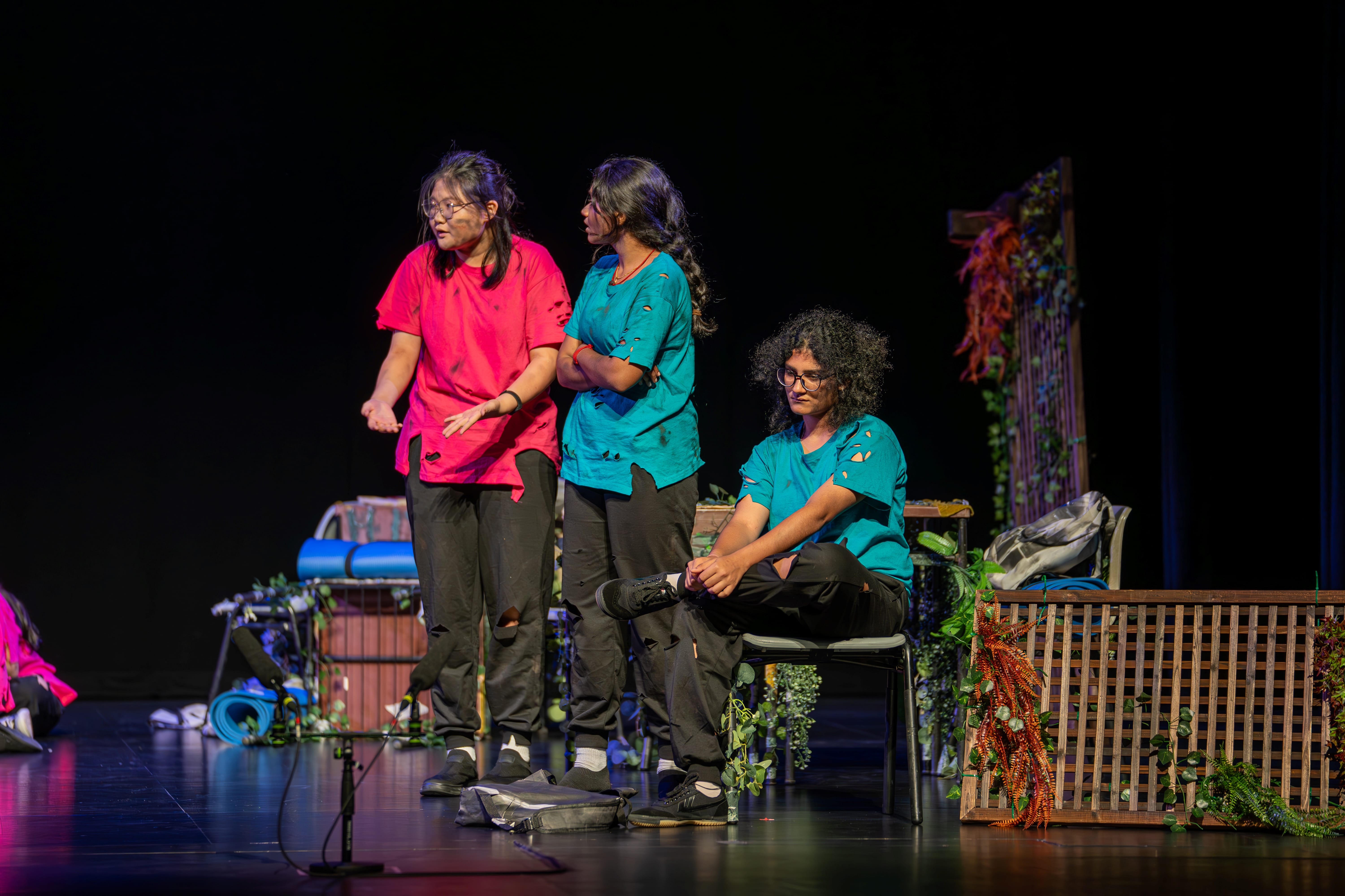 Three performers on stage; one in pink gestures, two in teal, with plants and props in background.