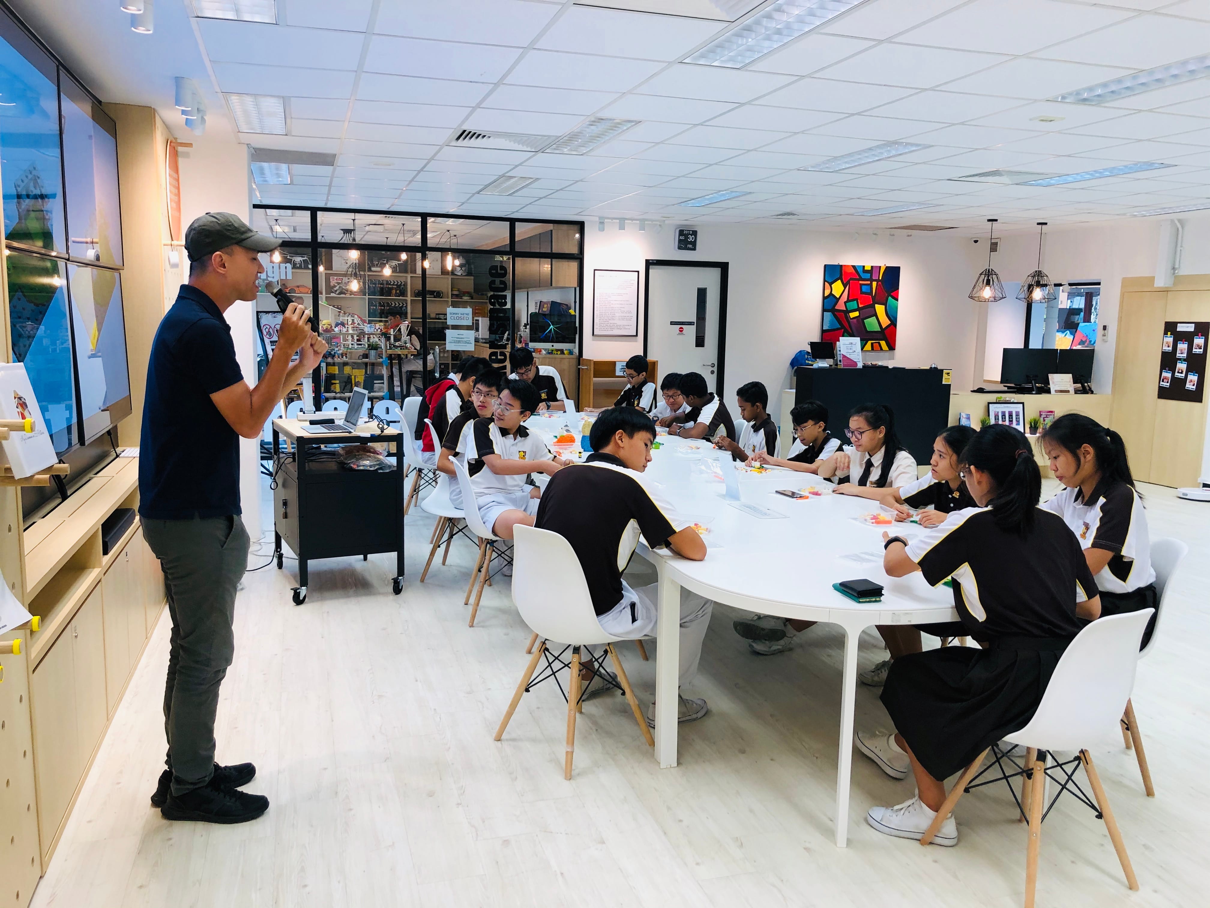 A man in a cap speaks to students in uniforms seated around a large white oval table in a bright classroom.