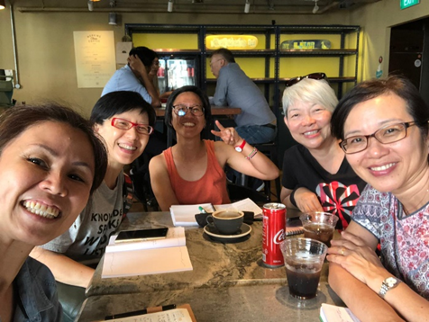 Five smiling women sit together at a cafe table with coffee and drinks, posing for a selfie.