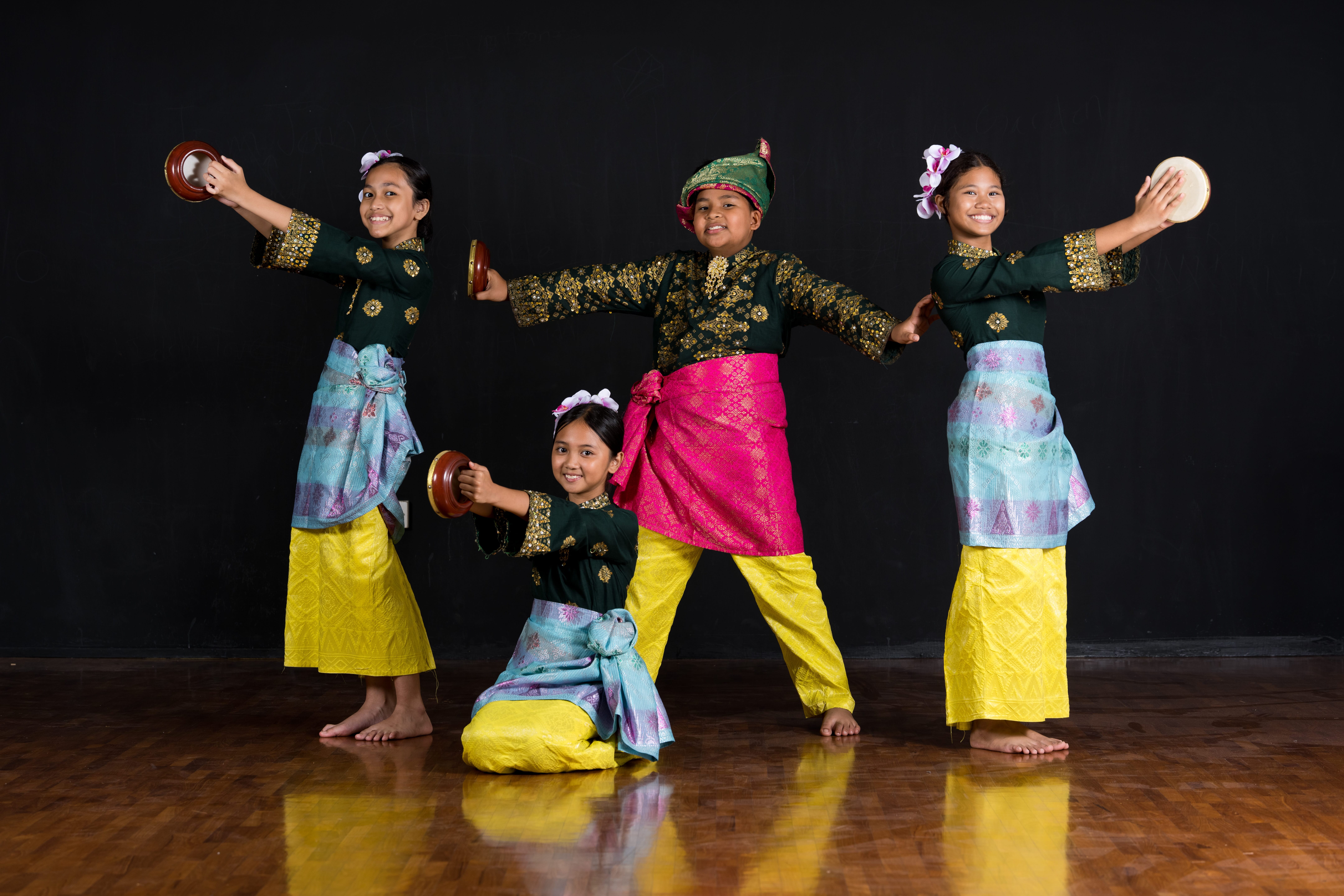 Four Evergreen Primary School students in colourful traditional Malay costumes pose with hand drums against a black backdrop.