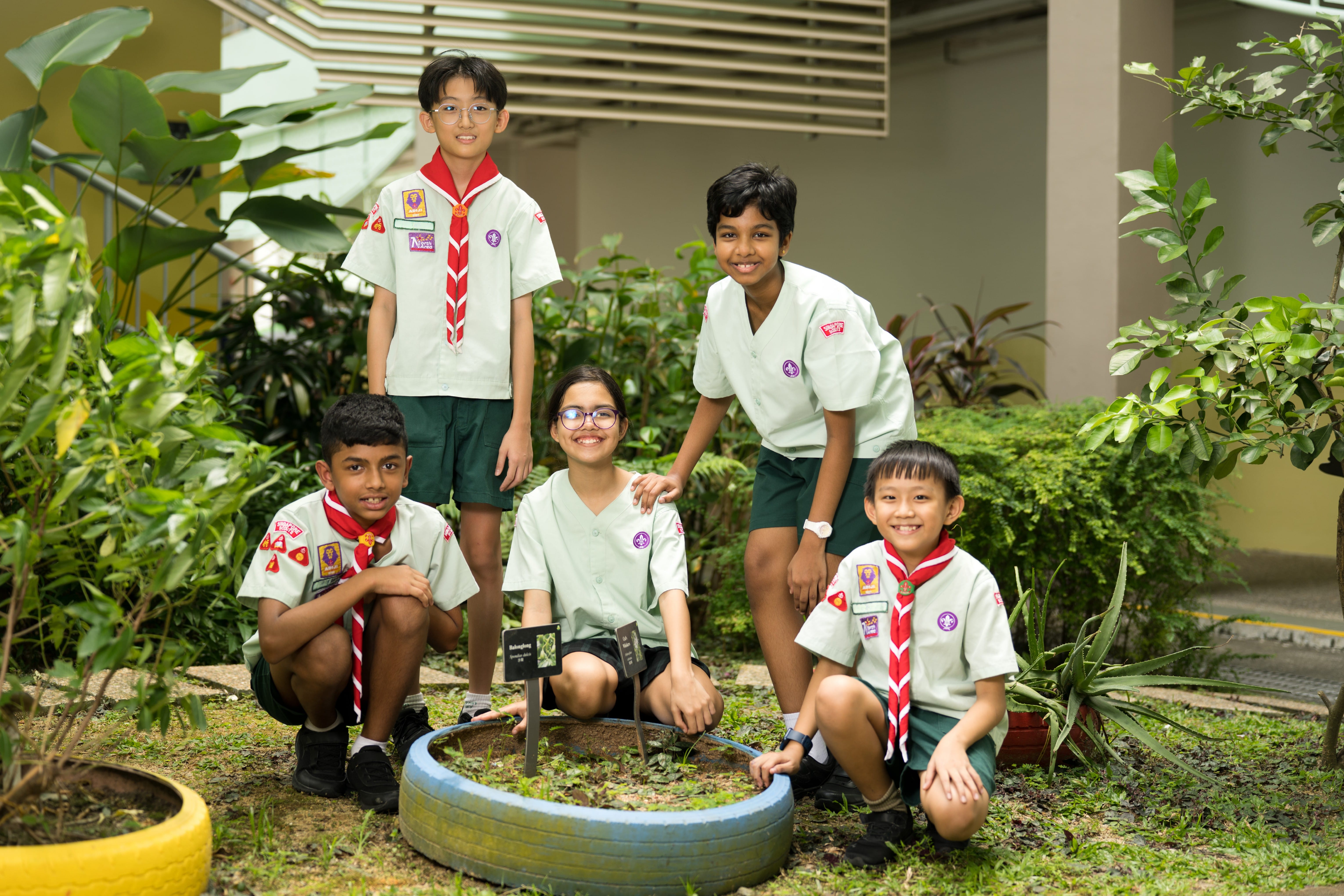Five children in scout uniforms smile together around a small garden planter made from a painted tire.