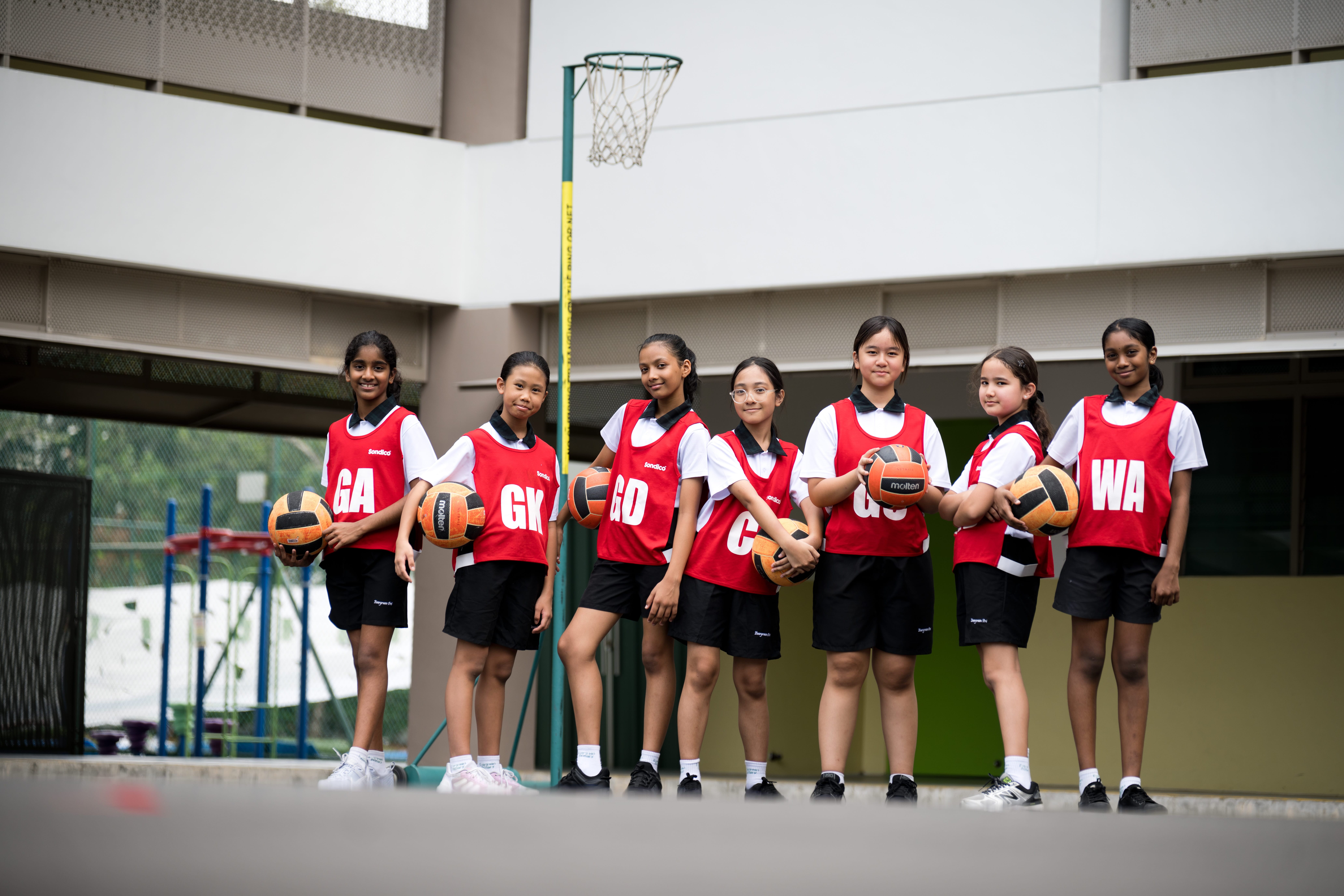 Seven Evergreen Primary School students wearing red netball bibs pose together on an outdoor court, each holding a netball.
