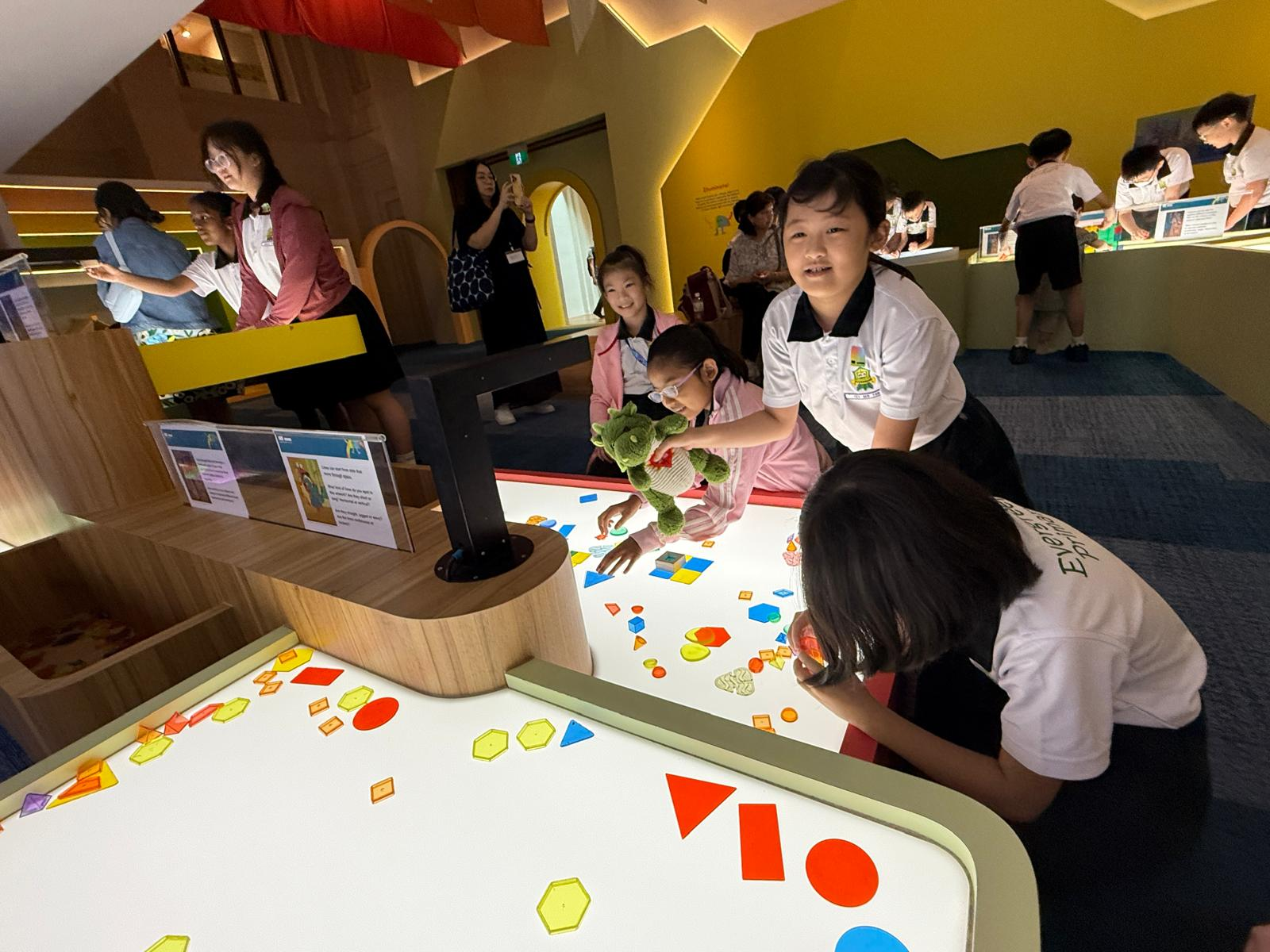 Students explore colourful geometric shapes on illuminated activity tables during an interactive learning experience, experimenting and working together through hands-on play.