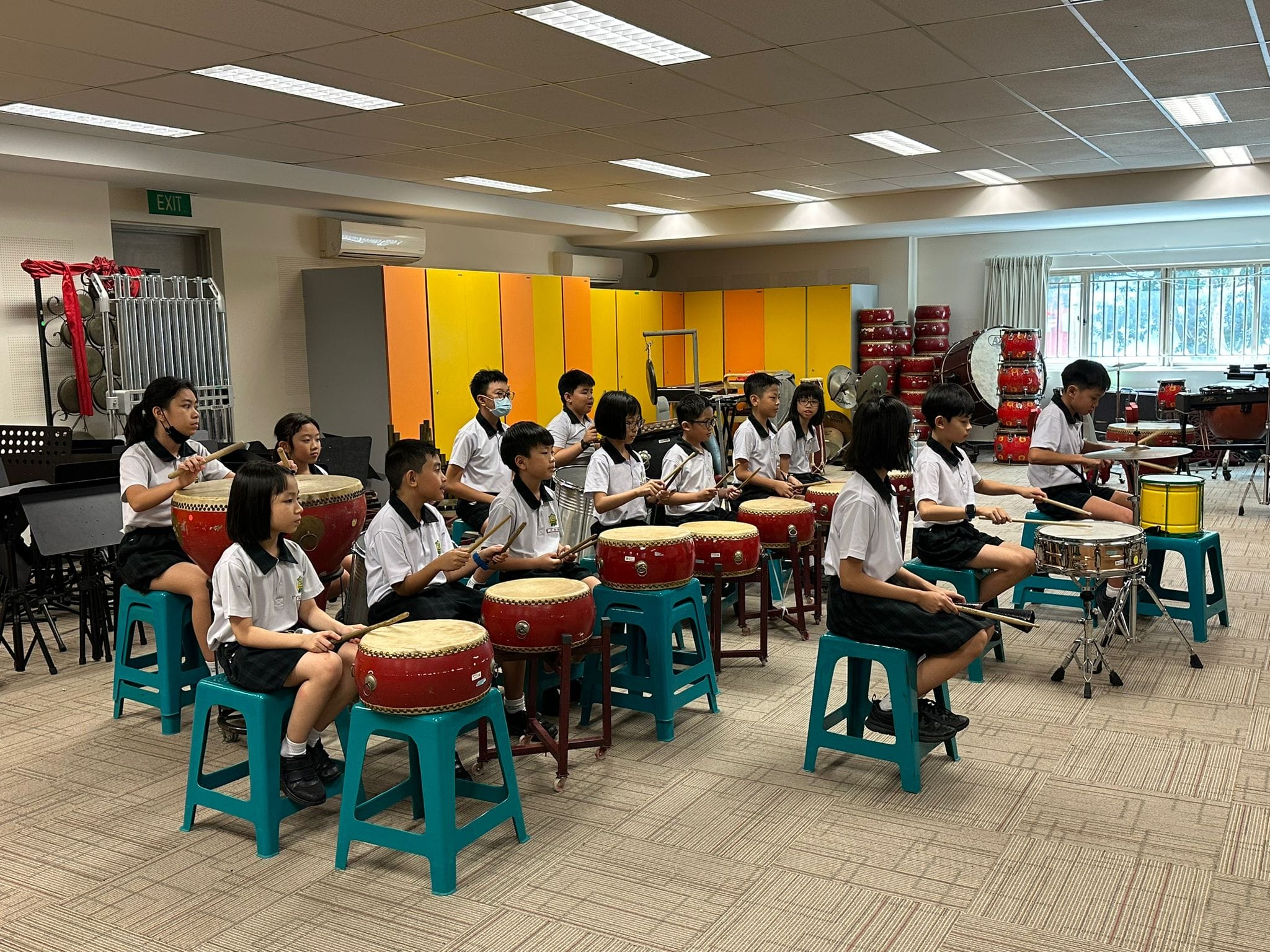 Students in school uniforms sit on teal stools playing red Chinese drums in a music room.