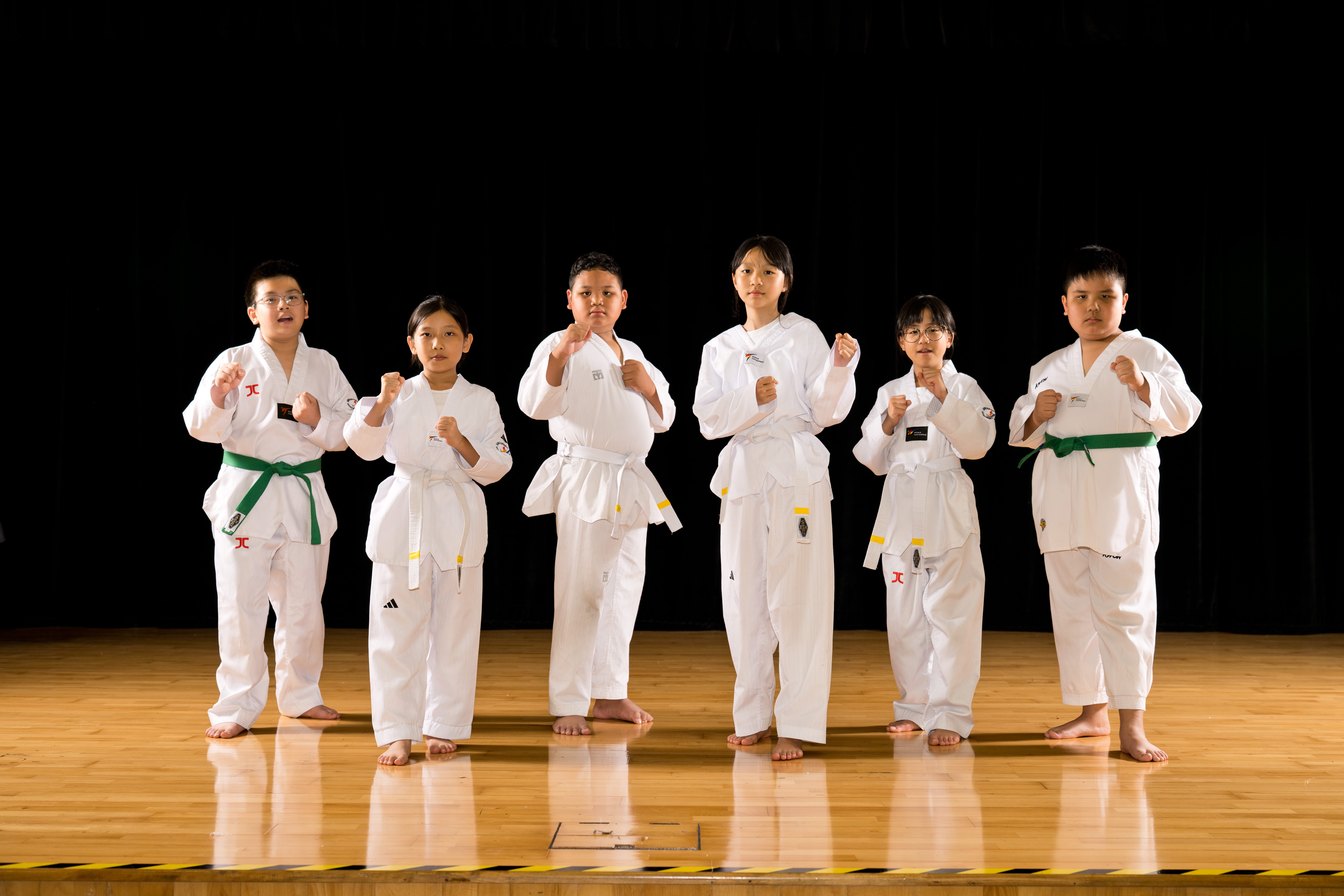 Six Evergreen Primary School students in white martial arts uniforms stand barefoot on a wooden stage against a black backdrop. Wearing different coloured belts, they pose confidently with their fists raised in ready stances.