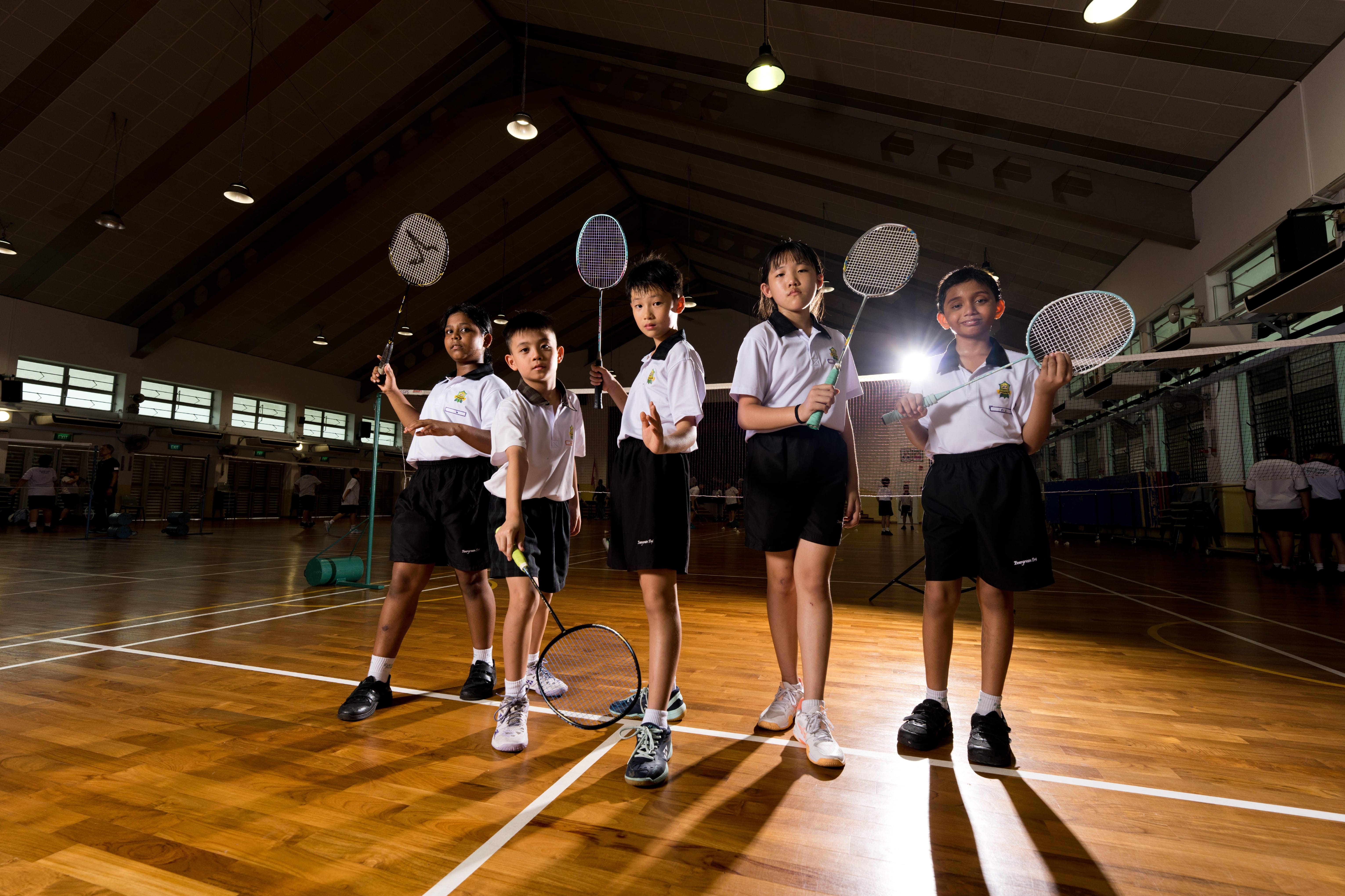 Five school children in PE attire hold badminton rackets on an indoor wooden court.