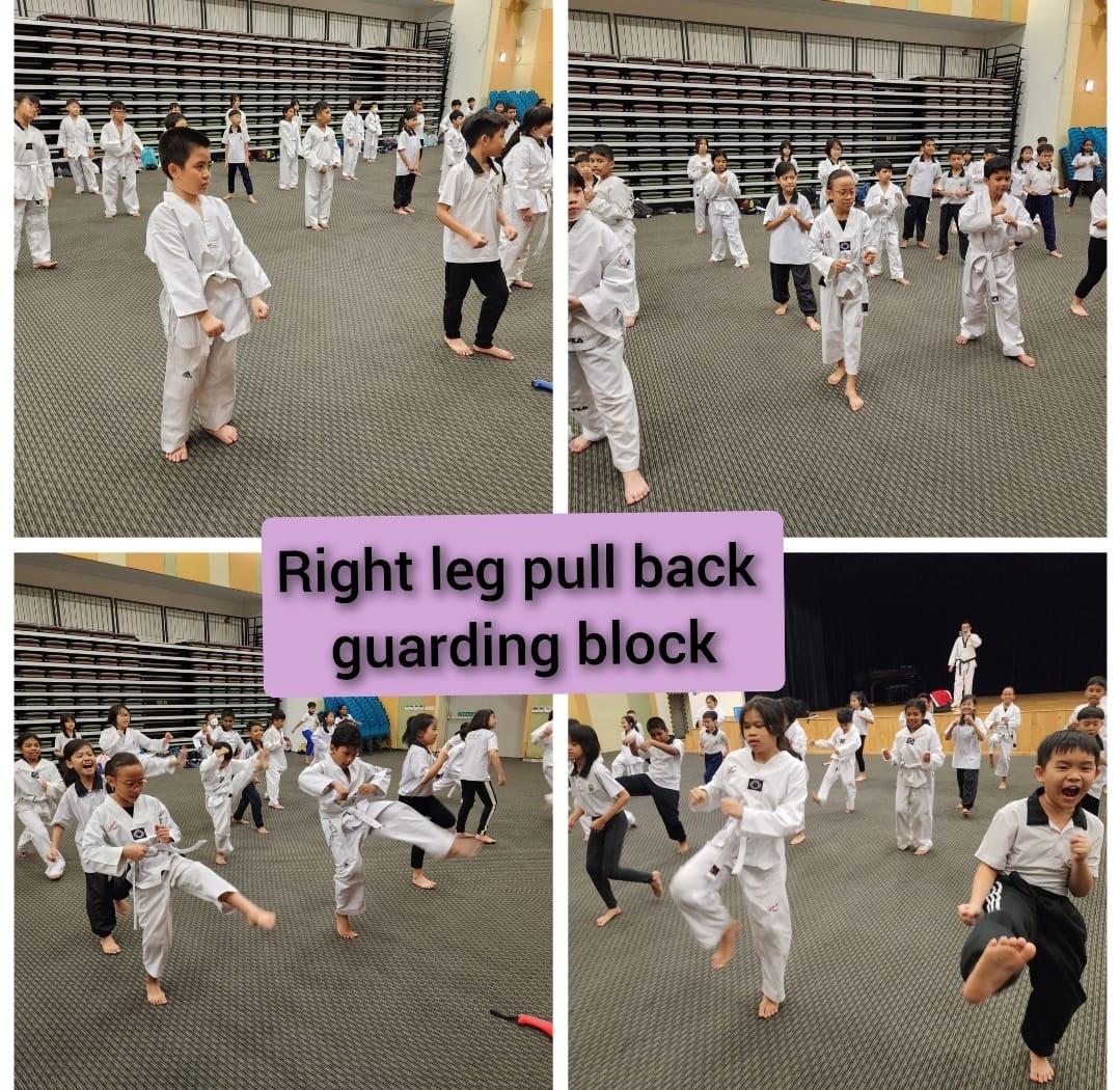Children in white taekwondo uniforms practice kicks and guarding blocks in a large indoor hall.