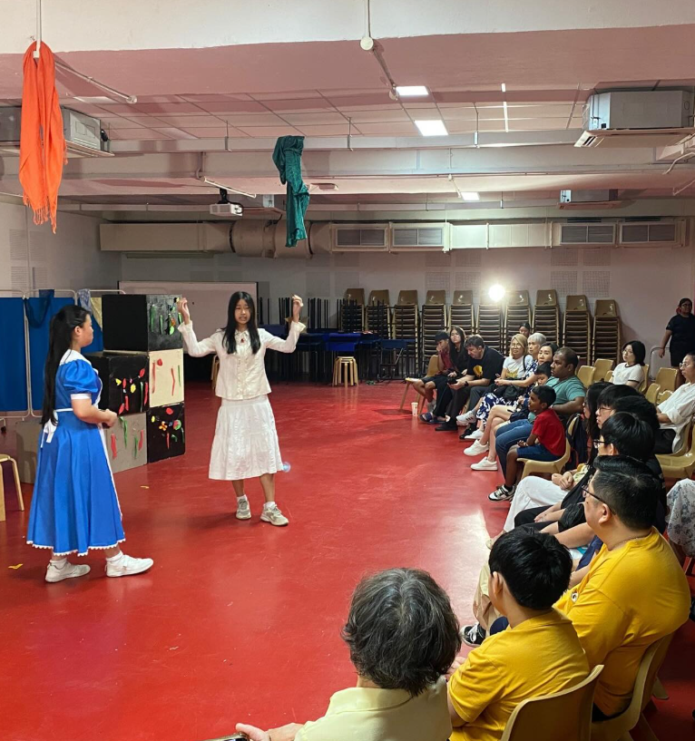 Two girls in costumes perform on a red floor for a seated audience in an indoor hall.