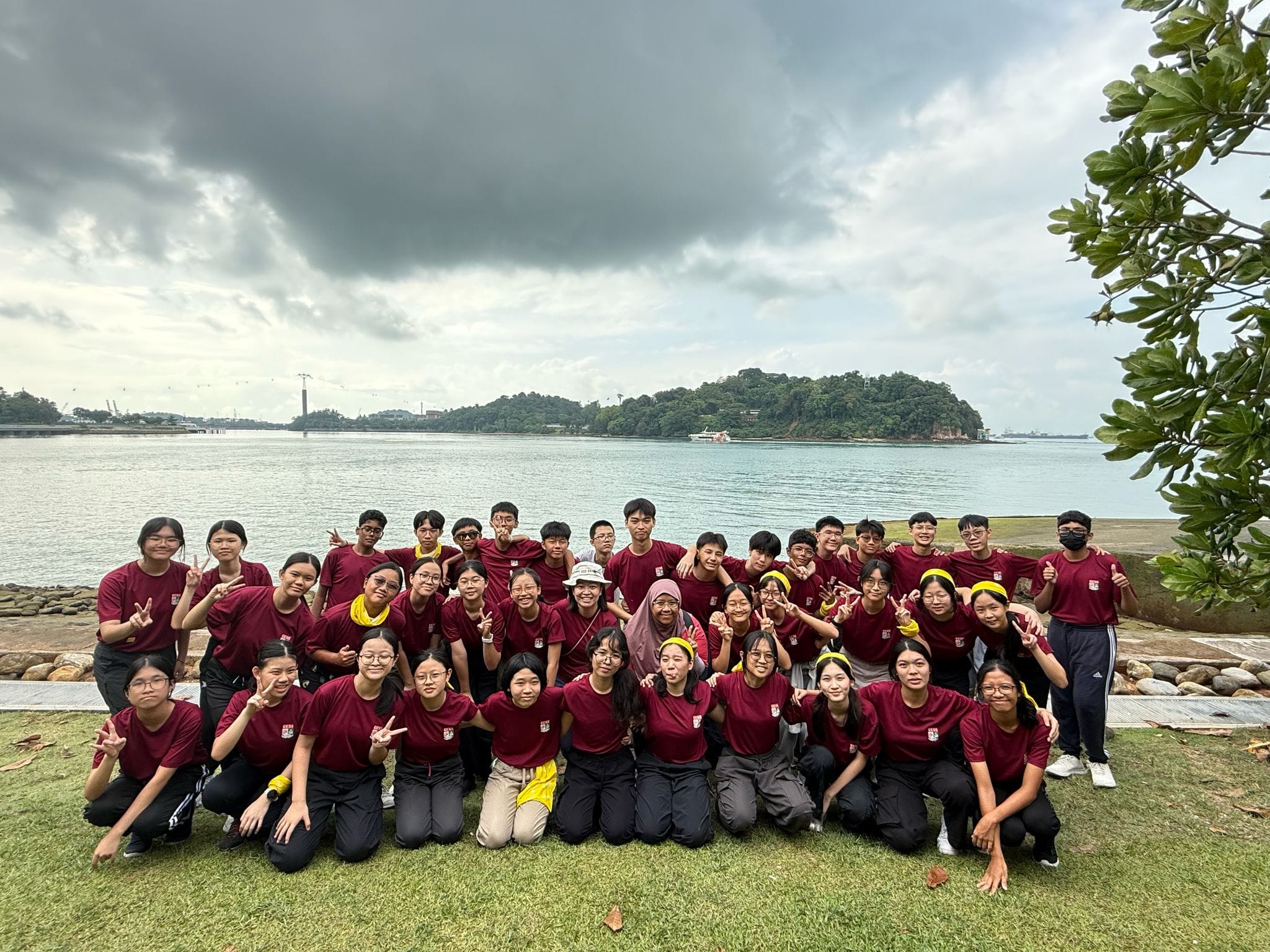 Large group of students in maroon shirts posing outdoors by a waterfront with a cloudy sky and green island behind them.