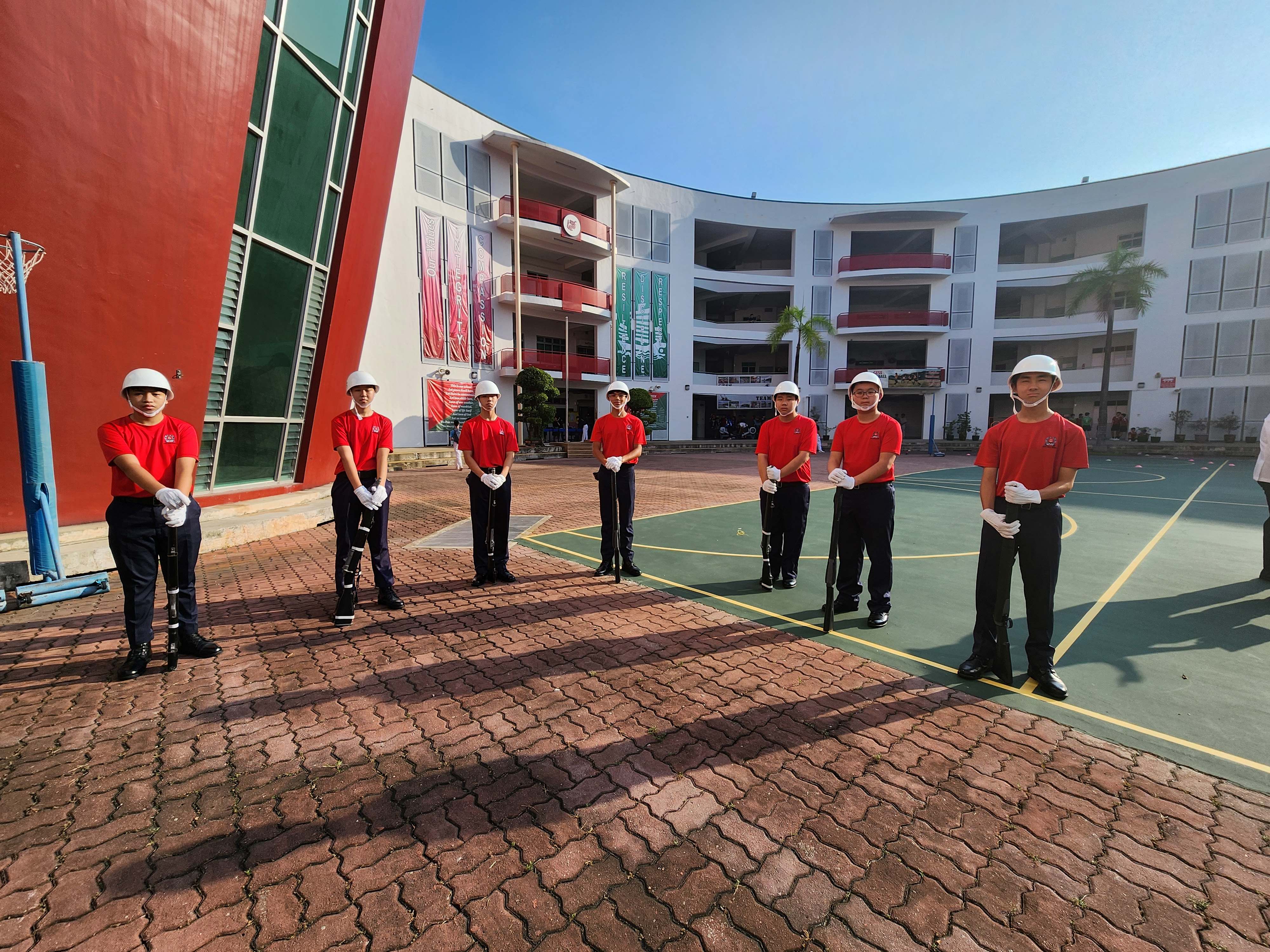 Seven youth cadets in red shirts, white helmets, and black pants stand at attention on a school courtyard.