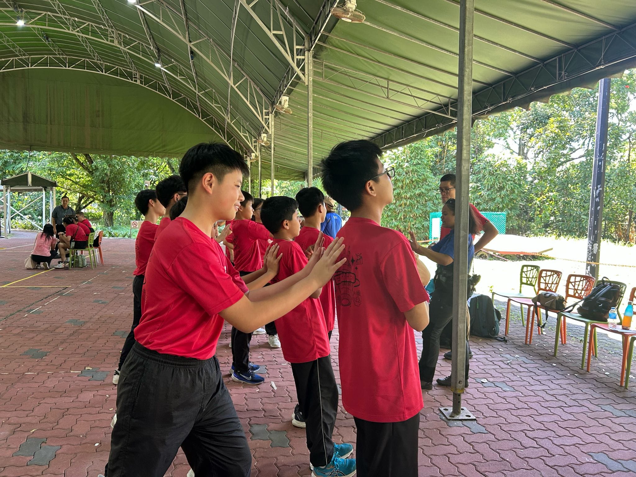 A group of young students in red shirts stand in a line under an outdoor canopy, with hands on each other's backs.