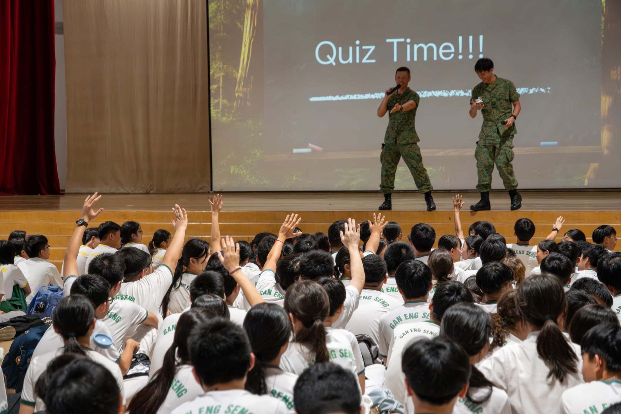 Students in white shirts raise hands during a Quiz Time session led by two soldiers on stage in a school hall.
