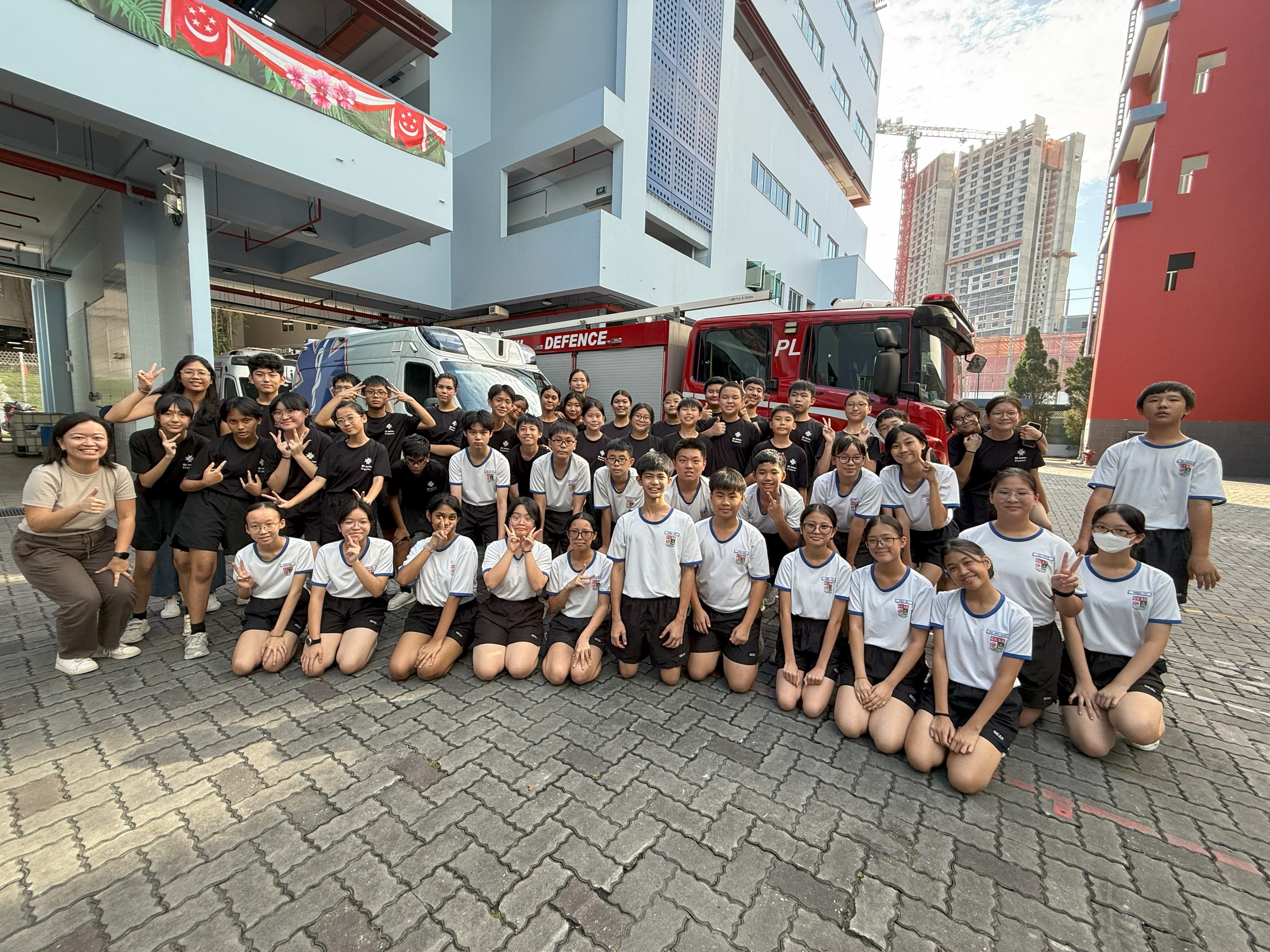 A large group of students in black and white uniforms pose outdoors in front of a red fire engine.