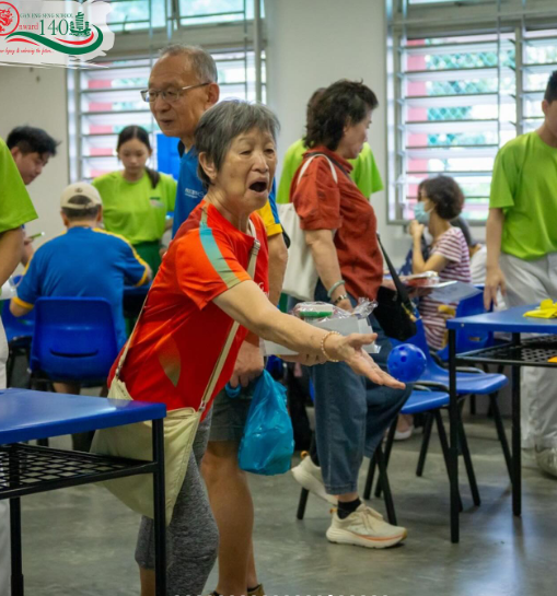 Elderly woman in red shirt gestures with open hand at an indoor community event with people in green and blue shirts.