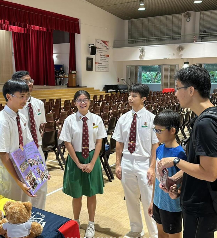 Students in white uniforms with red ties chat with a boy and an adult in a school hall with rows of chairs.