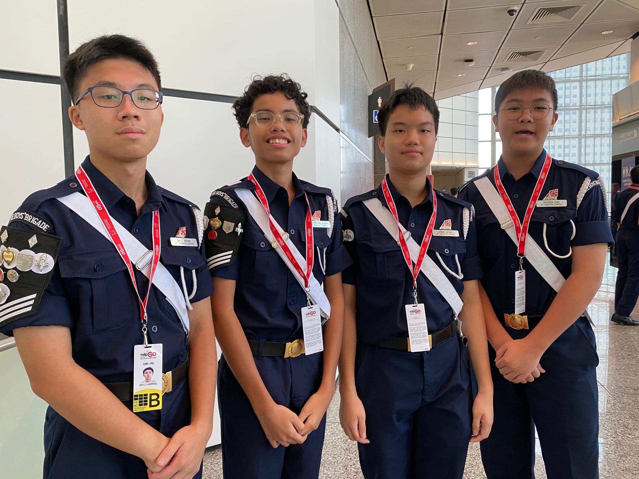Four boys in Boys Brigade navy uniforms with white sashes and red lanyards stand together indoors.