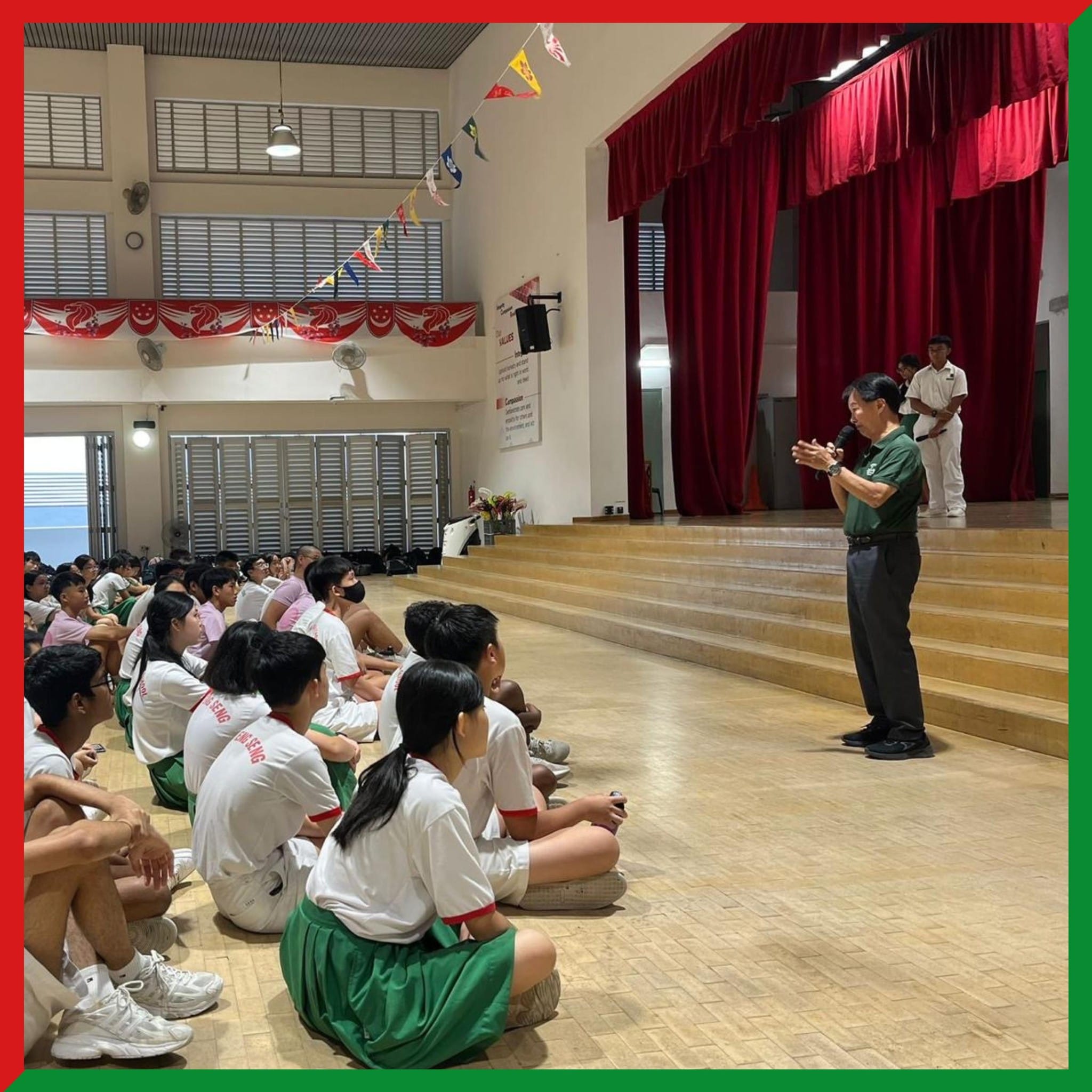 A man speaks into a microphone before students in white and green uniforms seated on a school hall floor.