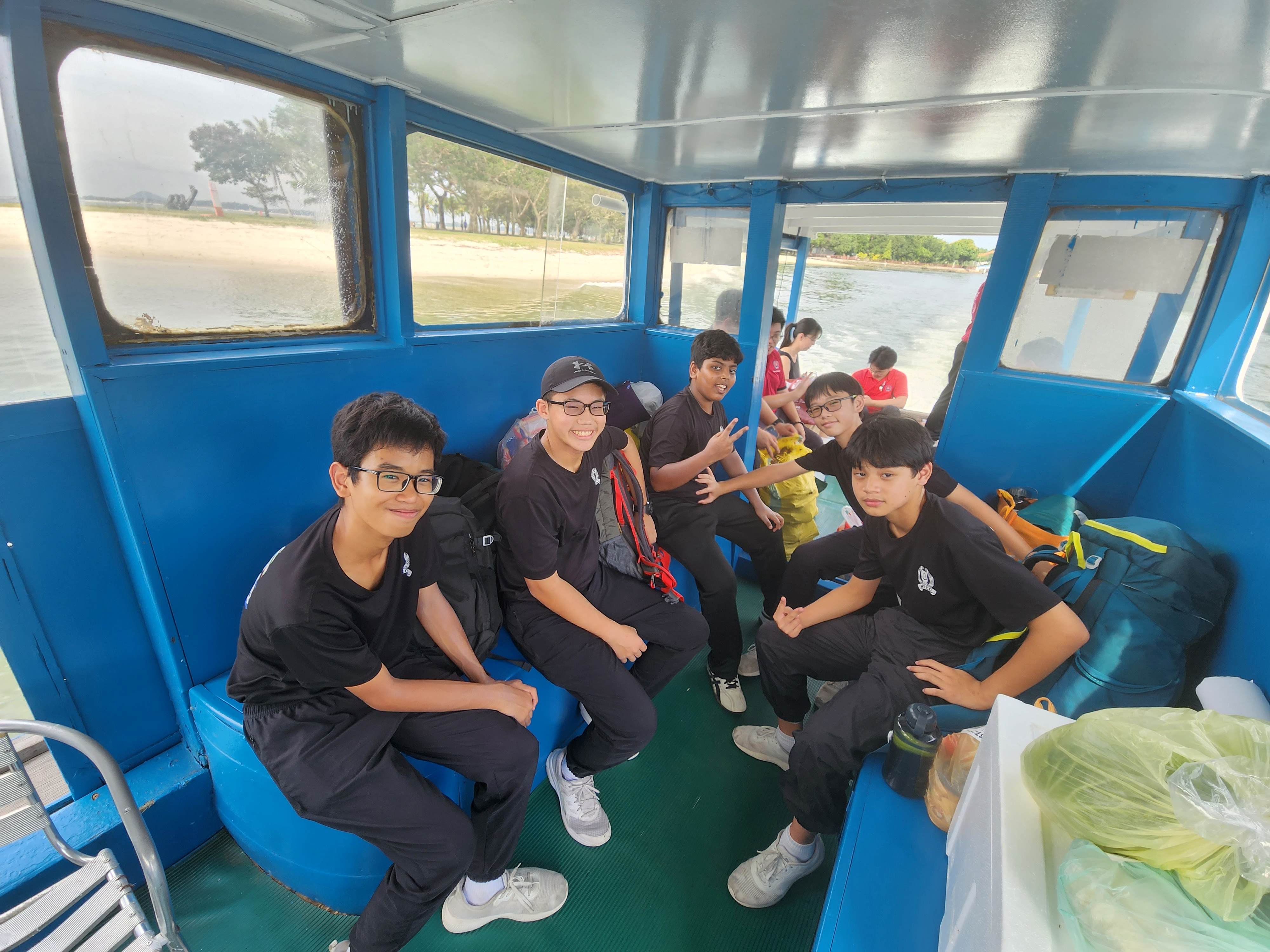 A group of young teens in black uniforms sit inside a blue-painted boat cabin, smiling at the camera.
