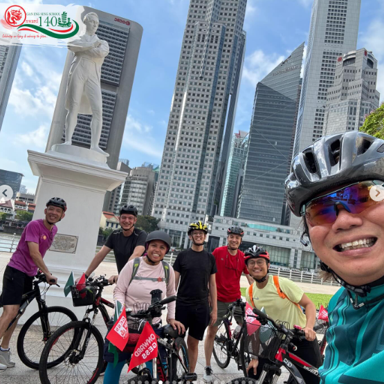 Six cyclists in helmets pose near a white statue with tall city skyscrapers in the background.