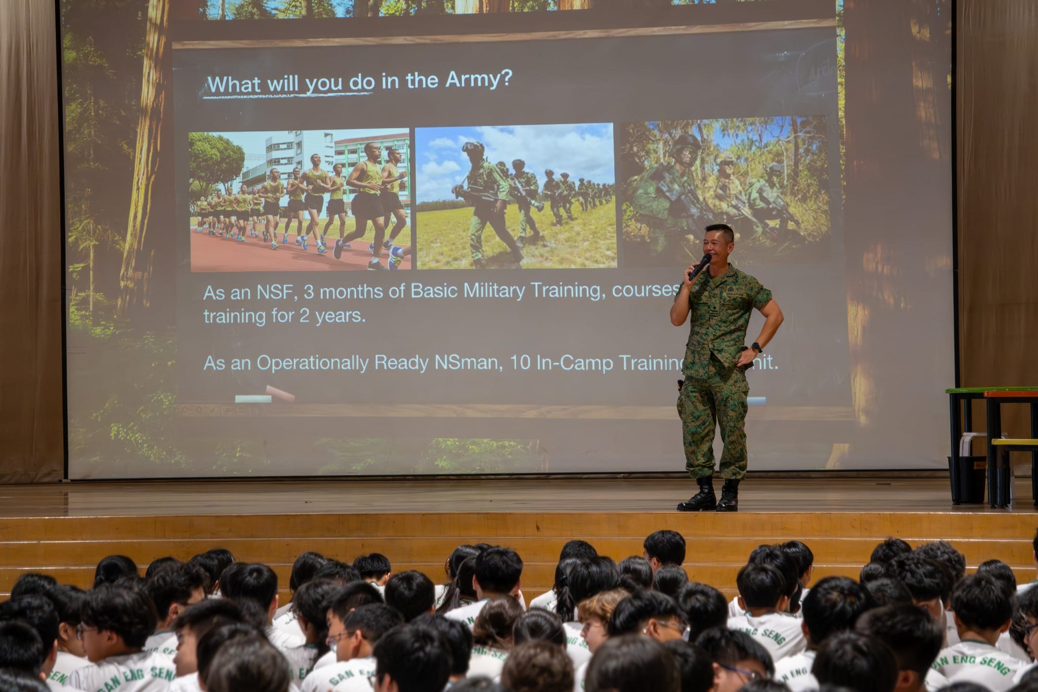 A soldier in camouflage uniform speaks on stage to a large seated student audience about army training activities.