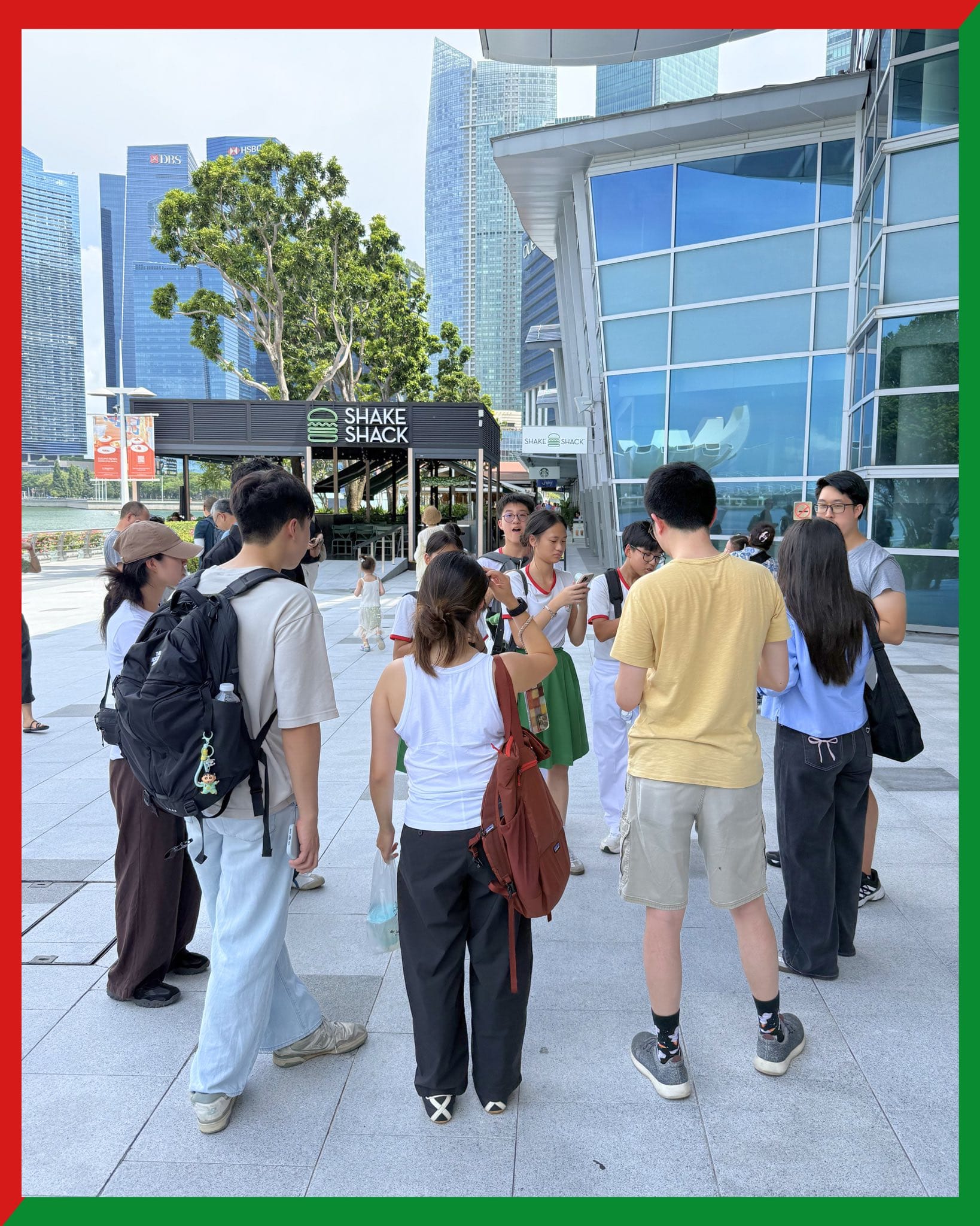 A group of people standing outdoors near a Shake Shack restaurant with tall city skyscrapers in the background.