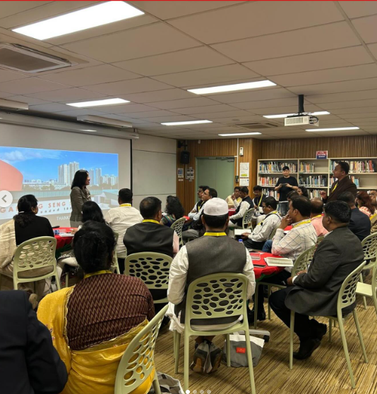 A presenter stands before a projected cityscape slide while attendees sit at tables in a library meeting room.