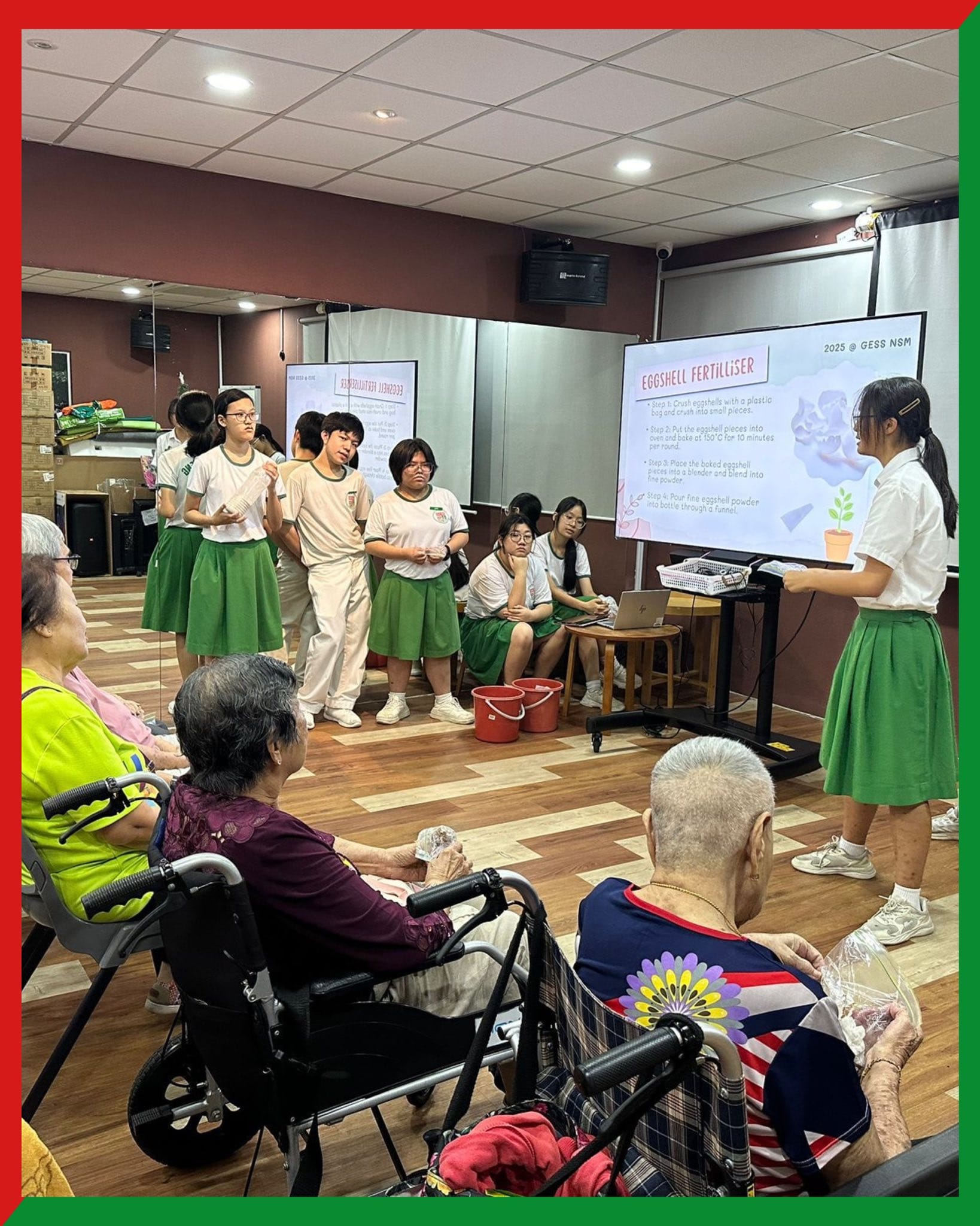 Students in white and green uniforms present an eggshell fertiliser slideshow to elderly seated audience members.