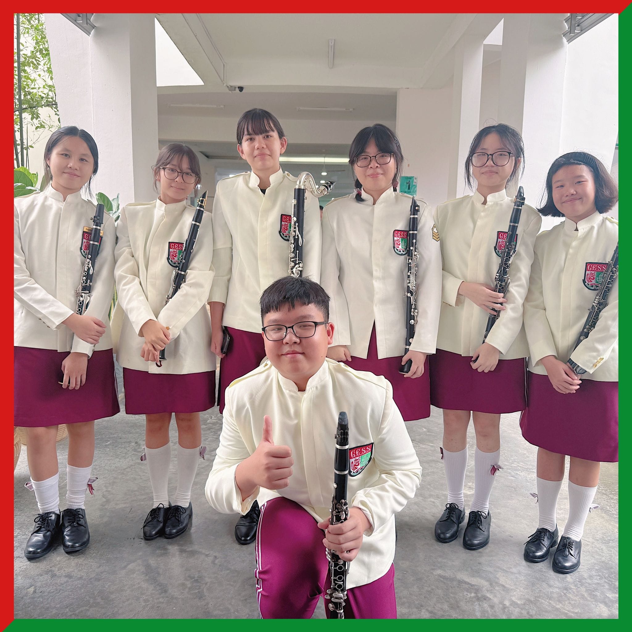 Seven students in white band uniforms holding clarinets pose together, one kneeling in front giving a thumbs up.