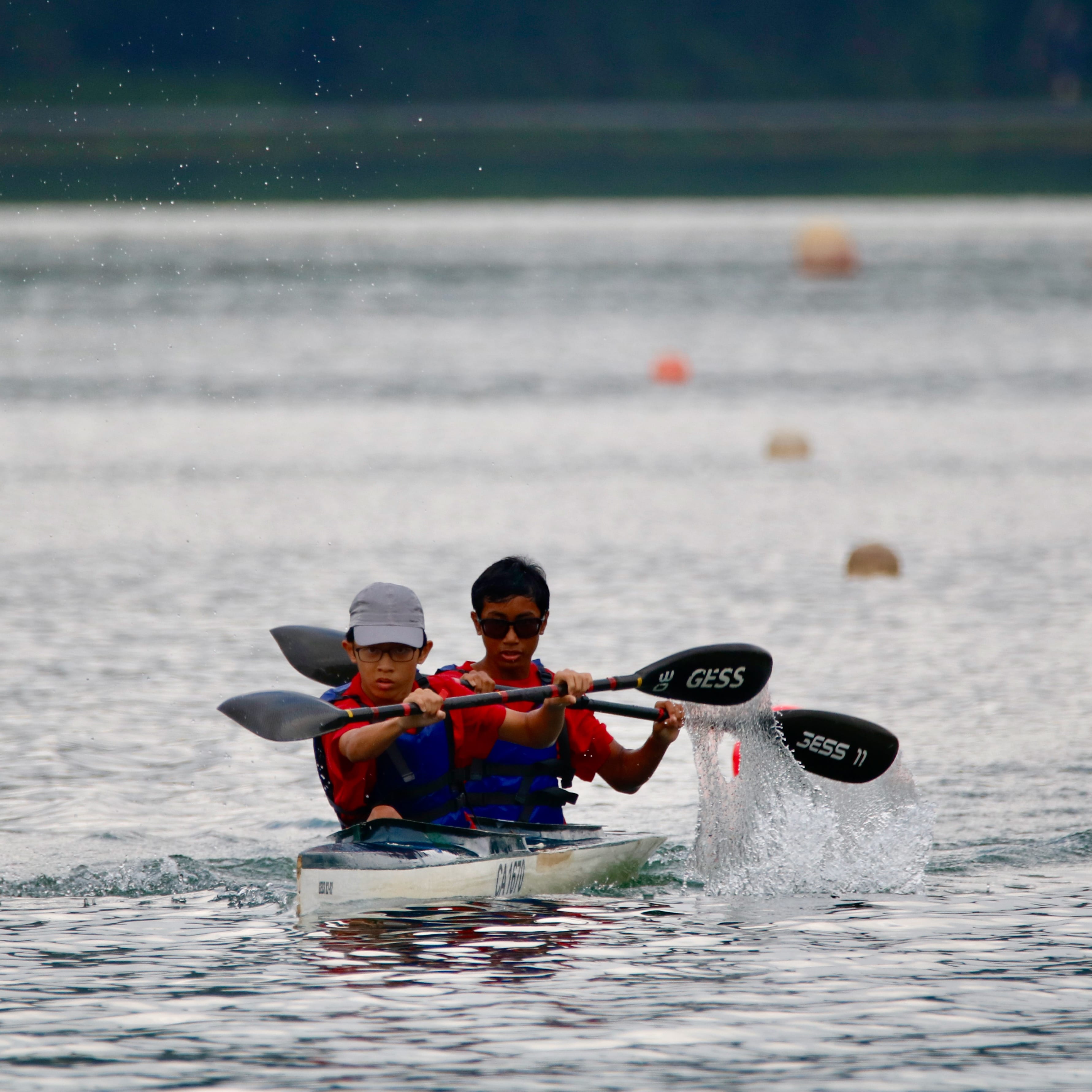Two young boys in red shirts and life jackets paddle a tandem kayak on open water with splashing oars.