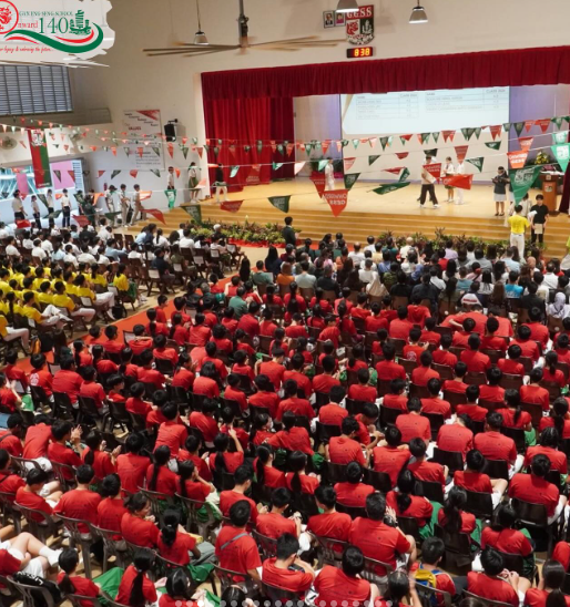 Large school assembly hall filled with students mostly wearing red uniforms, watching a stage event with speakers and a screen.
