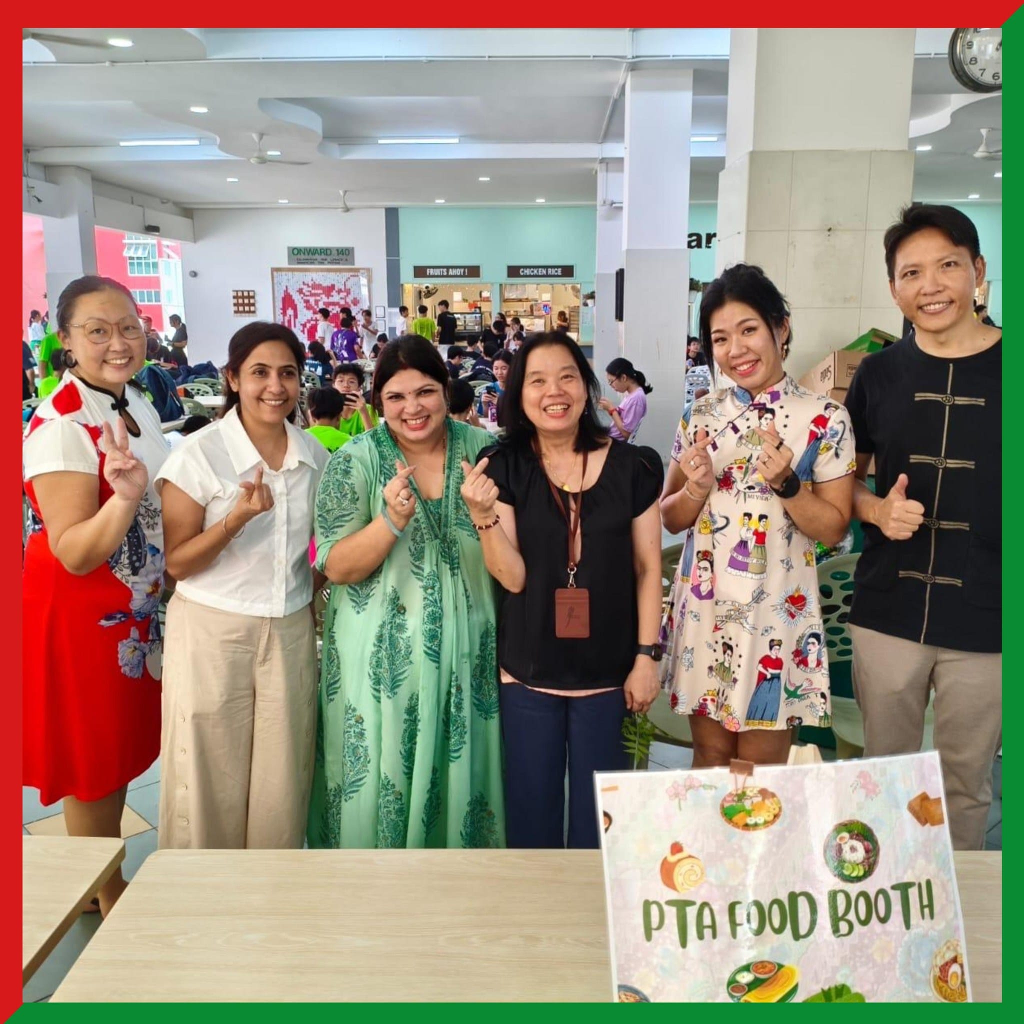 Six smiling women pose together at a PTA Food Booth sign in a busy school canteen.