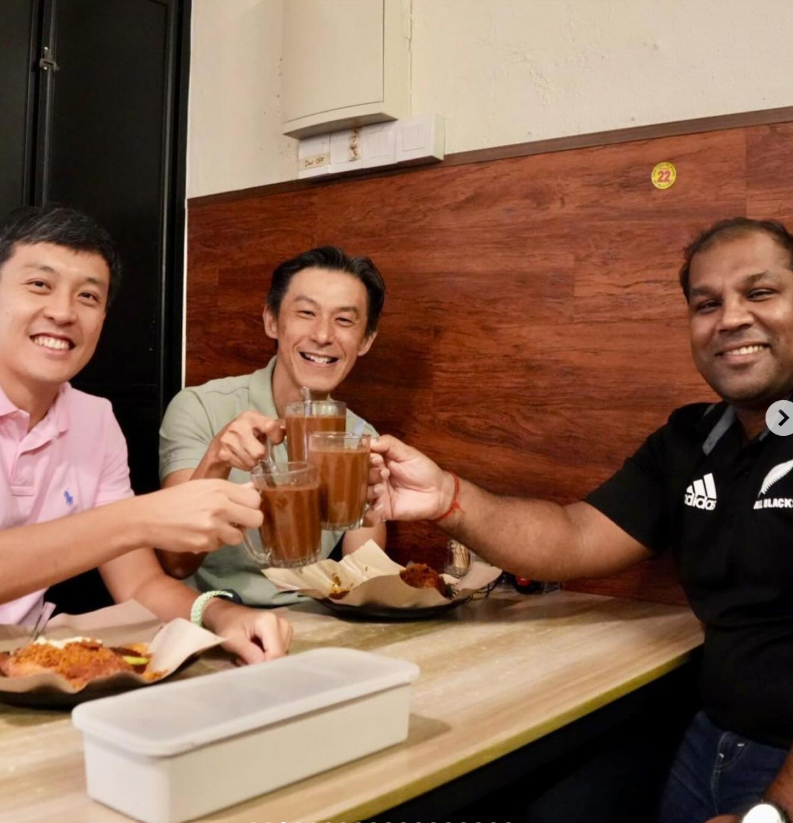 Three smiling men clink mugs of brown drinks together at a casual restaurant table with food nearby.