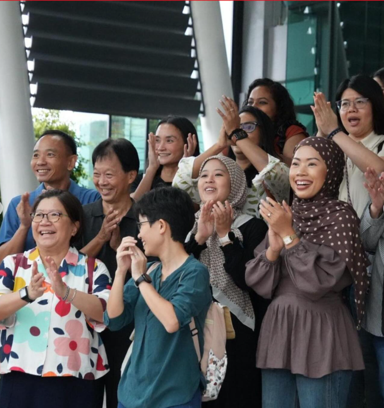 A diverse group of smiling people clapping and cheering outdoors near a modern building.
