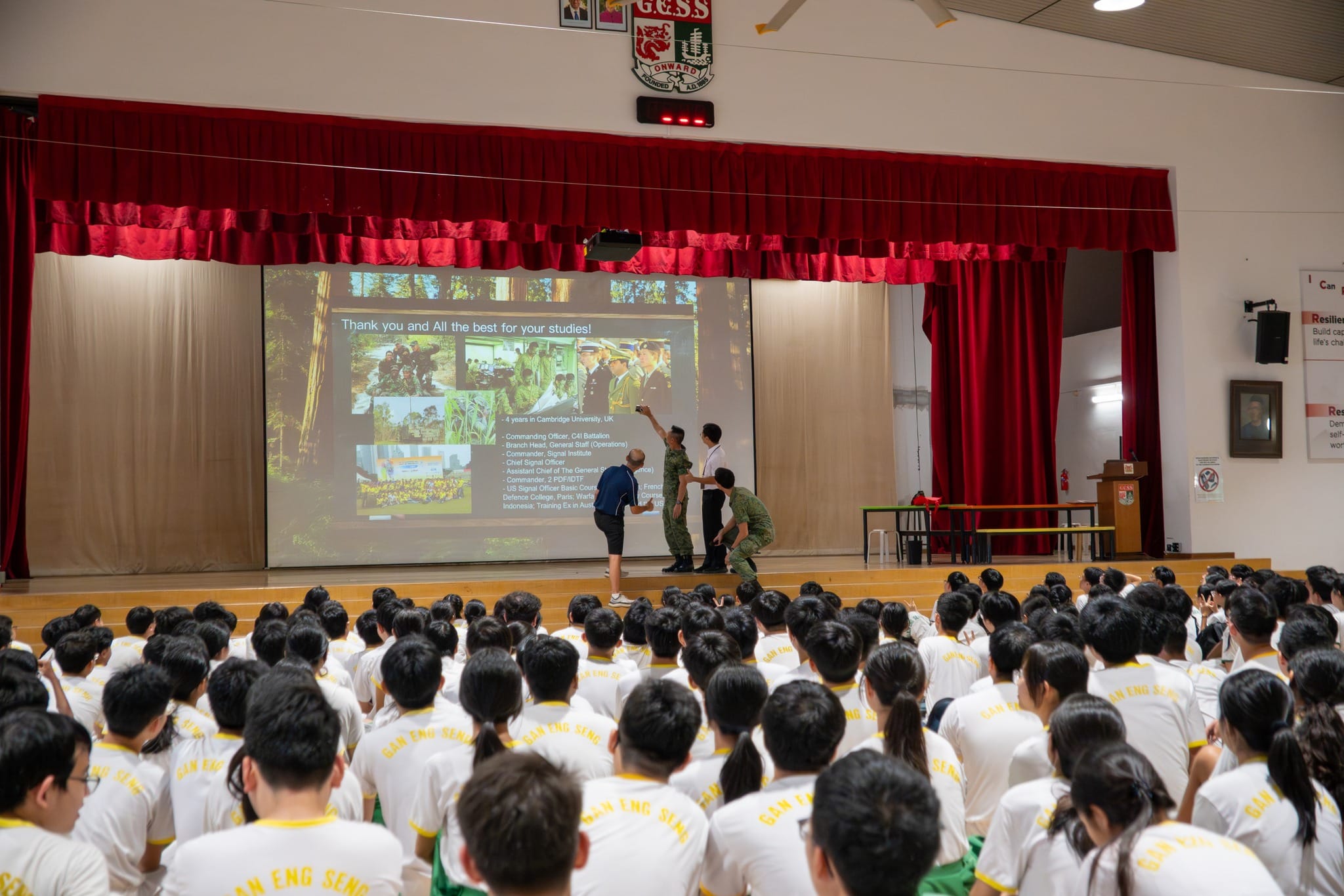 Students in white uniforms sit in a school hall watching a presentation slide on stage with speakers standing nearby.